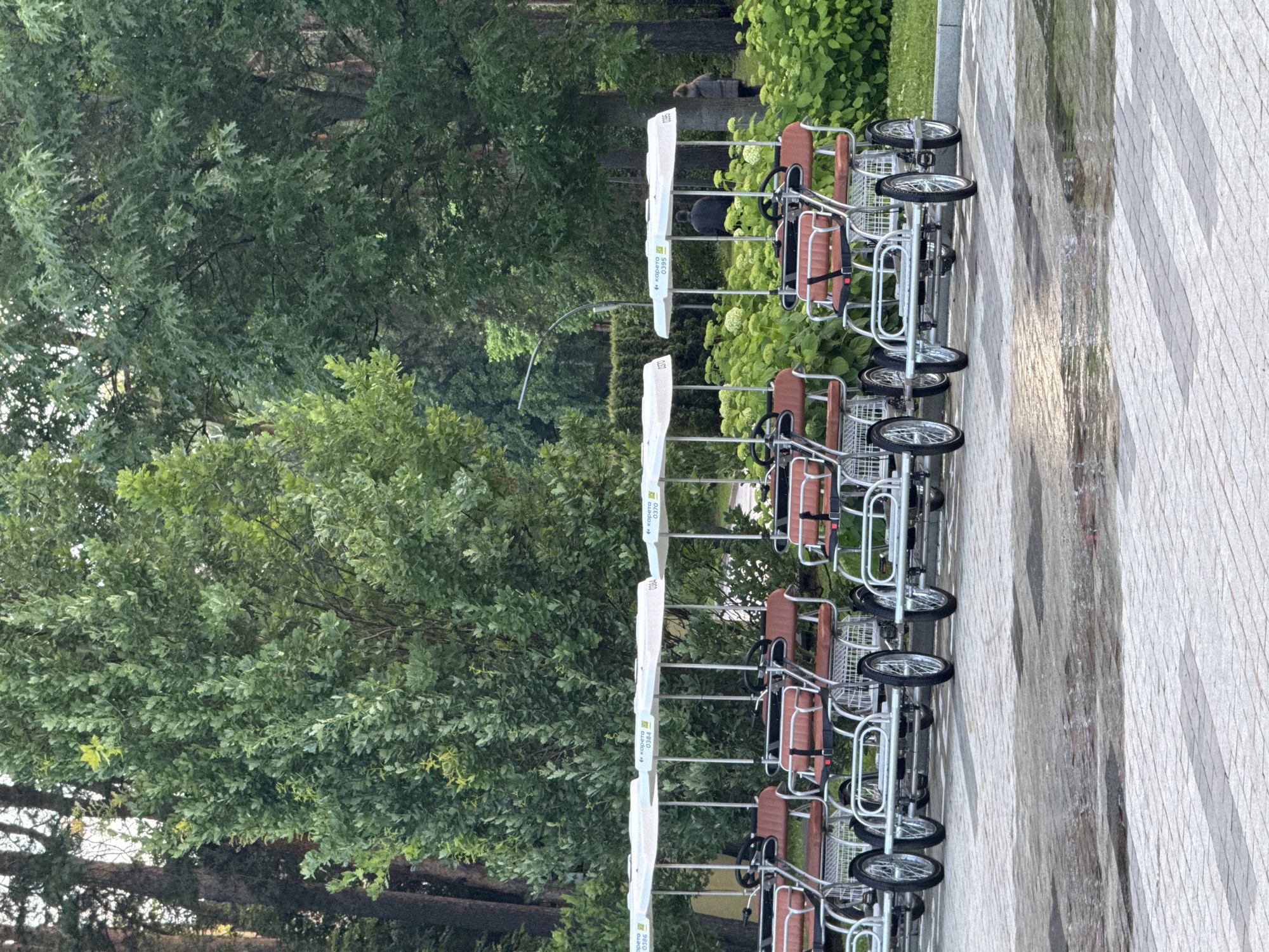 The image shows a row of multi-person pedal carts parked along a paved walkway in Moscow, Russia. The carts are white with brown seats and appear to be designed for recreational use. They are lined up facing to the right of the frame. The setting is outdoors, with lush green trees and foliage visible in the background. The ground beneath the carts is a light-colored paver with a diamond pattern, and parts of it appear wet, suggesting recent rain. The sky is overcast, indicating a cloudy day. One person, wearing a grey jacket, is visible in the distance, partially obscured by foliage. The scene suggests a quiet moment, possibly before or after a period of activity, with the carts waiting to be used. Visible text on the carts includes "Корета" (Koreta) and numbers such as "0370," "0384," and "0386," likely model or identification numbers. The overall mood is serene and tranquil.