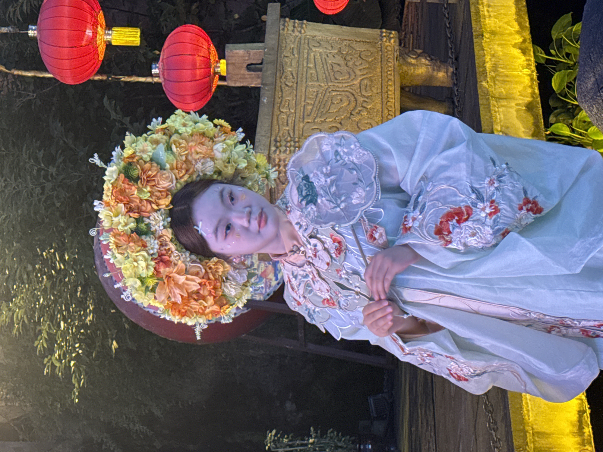 A young East Asian female, dressed in a light blue, elaborately embroidered traditional garment and a large, ornate floral headdress, is posed against a dark, outdoor background with hints of trees and foliage. Red Chinese lanterns are visible, hanging from strings, and a golden, carved urn-like object is positioned behind her. The female has white and pink face paint, resembling flowers, on her cheeks and forehead. Her hands are clasped together in front of her, holding what appear to be thin sticks. The lighting suggests it is nighttime or dusk, with a bright yellow illuminated platform in the foreground. The location is Yuzhong District, China.