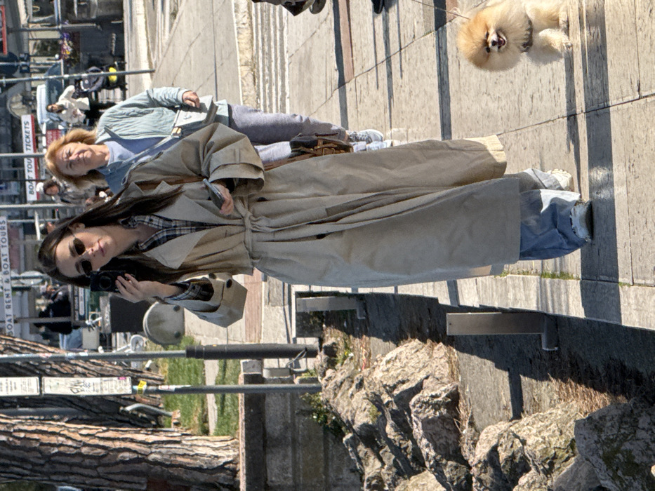 The scene depicts a sunny daytime view of a paved promenade in Peschiera del Garda, Italy, likely alongside a waterfront.
In the immediate foreground, a young adult female with long dark hair, wearing sunglasses, a plaid shirt, a long beige trench coat tied at the waist, loose blue jeans, and white sneakers, walks towards the viewer. She holds a small black electronic device to her right ear/face and a smartphone in her left hand. A small brown shoulder bag is slung over her left shoulder. To her right, a small, fluffy, light orange/cream-colored Pomeranian-type dog on a leash walks on the paved surface, appearing to smile.
Behind the primary subject, an older adult female with light reddish-brown hair, wearing a light blue quilted jacket, grey sweatpants, and white sneakers, walks in the same direction, carrying a light-colored crossbody bag.
To the left, a low concrete wall separates the promenade from a rocky embankment. A large tree with rough bark is visible on the far left, behind the embankment. Several short, L-shaped grey bollards are positioned along the edge of the paved path.
In the background, across a street or road, multiple signs are visible, including "BOAT RENT & BOAT TOURS" and "BOAT TOUR TICKETS," indicating proximity to a lake or harbor. Distant blurred figures and vehicles are also present. Strong sunlight from the right casts clear shadows on the walkway.