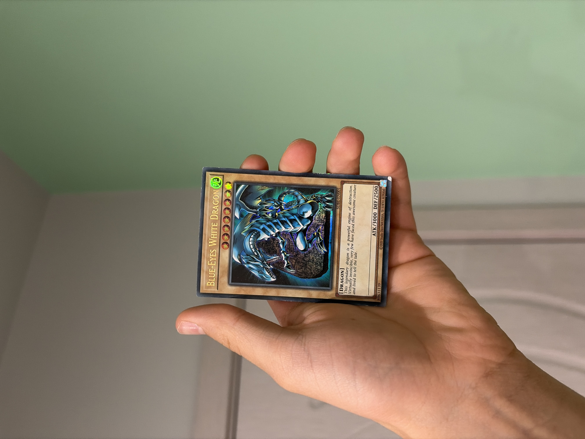 A person's hand is holding a "Blue-Eyes White Dragon" Yu-Gi-Oh! card. The card is positioned in the center of the frame, with the person's thumb and fingers supporting it. The background is out of focus, showing a light-colored wall and a section of a pale green wall. There is no discernible location information to suggest the city of Svetovrachene, Bulgaria.