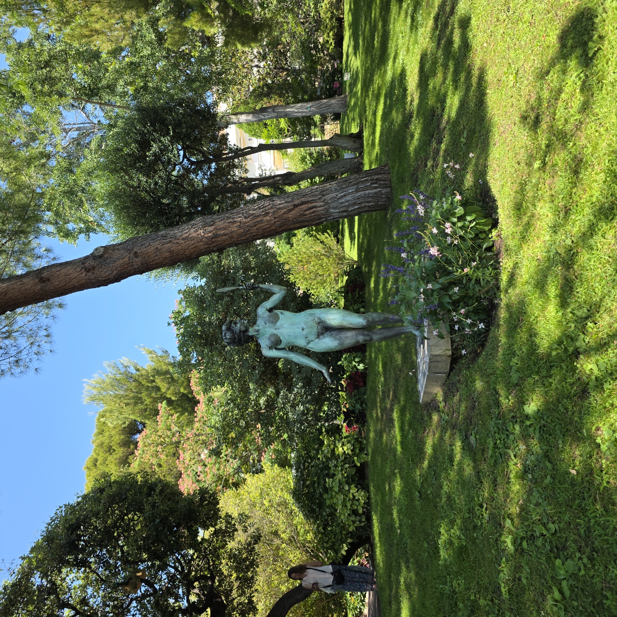 In Monaco-Ville, Monaco, within the Japanese Garden, a bronze statue of a nude female figure stands on a stone pedestal. The statue, weathered to a greenish patina, depicts a woman holding a handheld mirror in her right hand, with her left arm extended downwards. She is positioned amidst lush greenery, with a large tree trunk to her right and various plants and flowers surrounding the base of her pedestal. In the background, a woman in a light-colored top and patterned skirt is visible, standing to the left near a curved tree. The garden features manicured green lawns, dense foliage, and a clear blue sky. The sunlight casts dappled shadows across the grass, indicating a bright day.