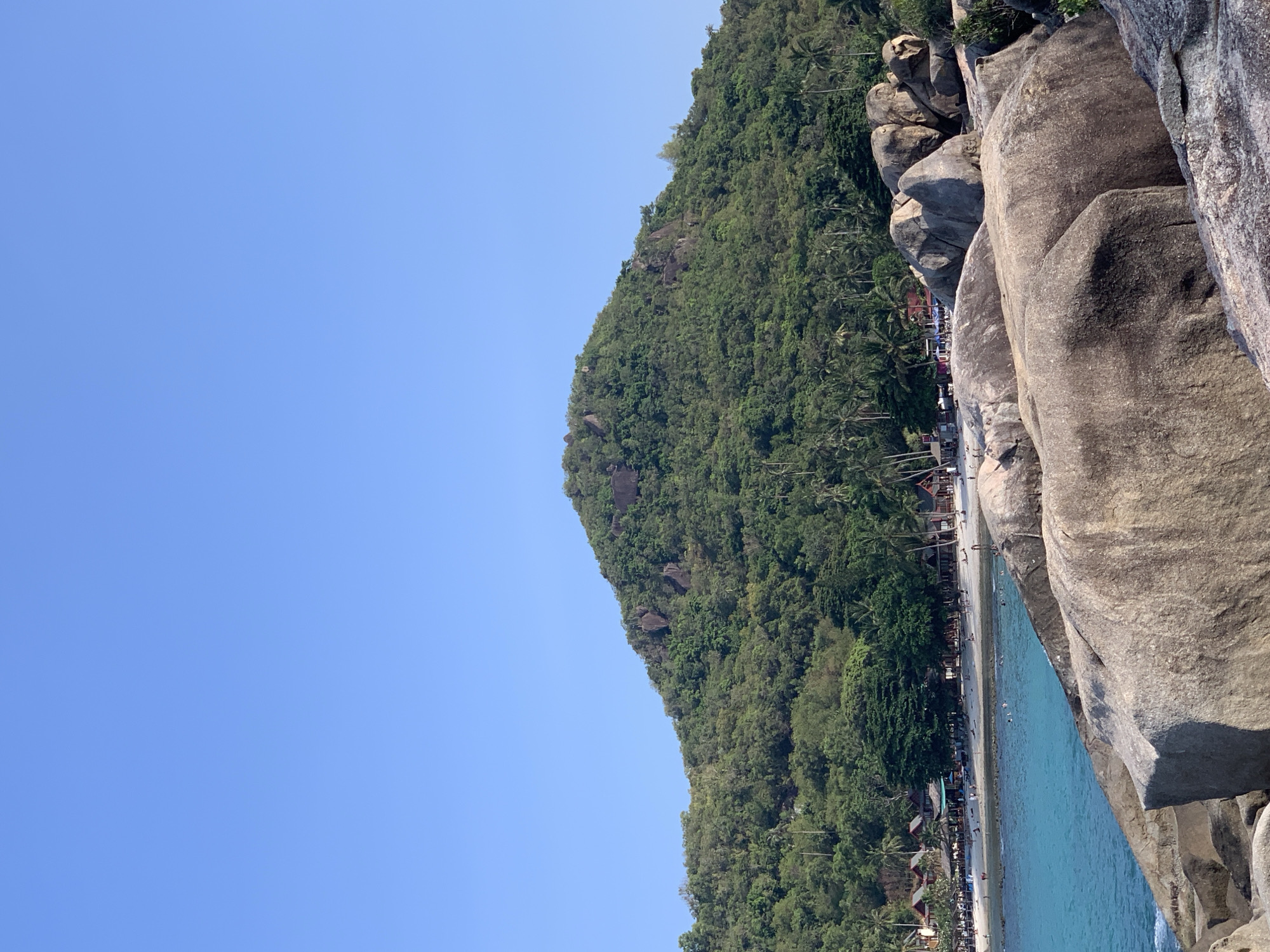 The image captures a coastal scene near Baan Chaweng Noi, Thailand. Dominating the right side are large, grey, weathered rocks, providing a rough, textured foreground. Adjacent to the rocks is a beach with numerous small buildings, possibly resorts or residences, along the tree line. A crowd of people populate the beach and the water. The water appears turquoise and calm, reflecting the clear sky. Beyond the beach, a densely vegetated hillside rises, covered in lush green trees and occasional rocky outcrops. The sky is a clear, bright blue, taking up the left side of the frame. The scene portrays a sunny day at a tropical beach resort.