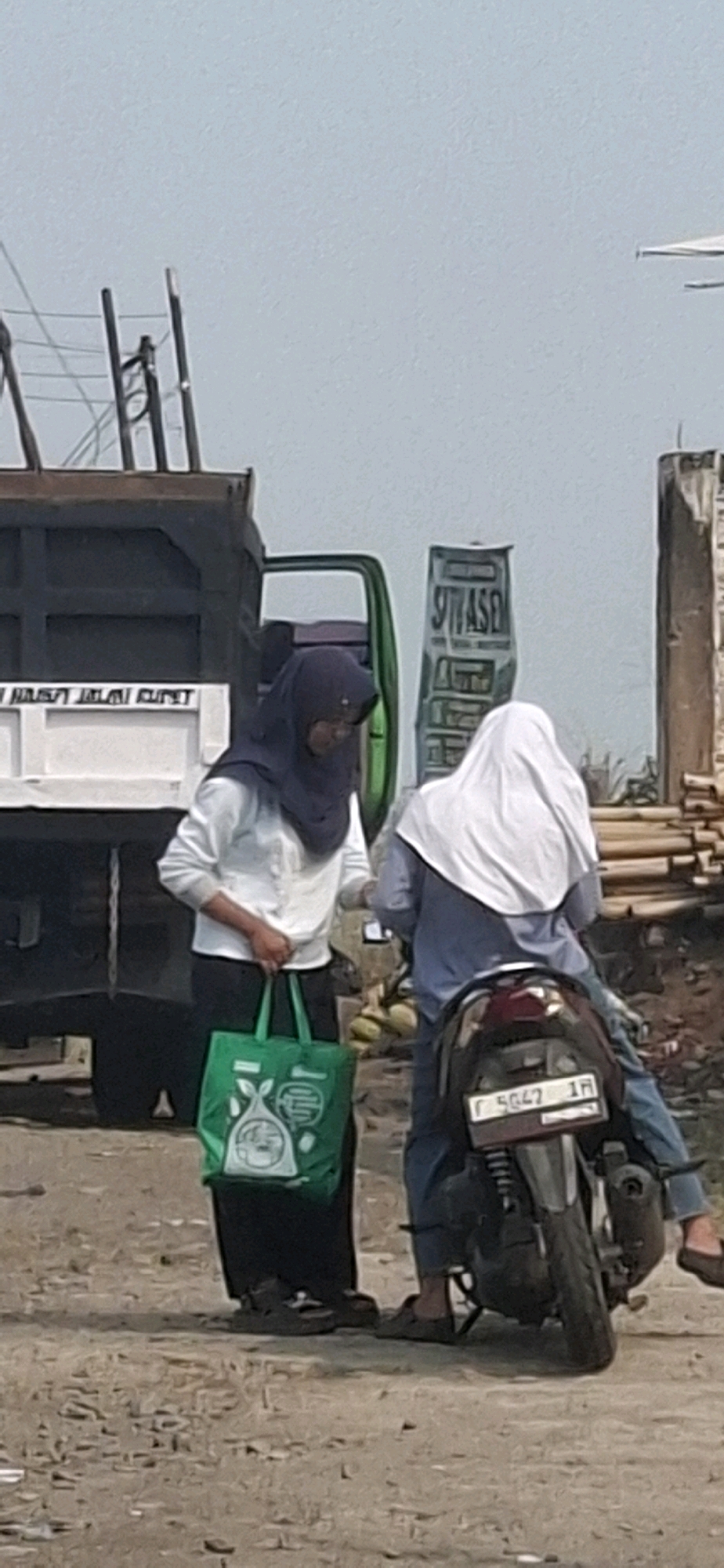 In the city of Cibinong Hilir, Indonesia, two women are interacting outdoors. One woman, wearing a dark blue hijab and a white long-sleeved shirt with black pants, stands on the left, holding a green reusable shopping bag. The other woman, wearing a white hijab and a blue long-sleeved shirt with blue jeans, is seated on a dark-colored motorcycle. The motorcycle has a license plate that reads "F 5647 1H". Behind the women, a large dark green truck is visible, and to the right, stacks of lumber and a partially constructed building are present. The ground is covered in dirt and gravel. The sky is overcast.