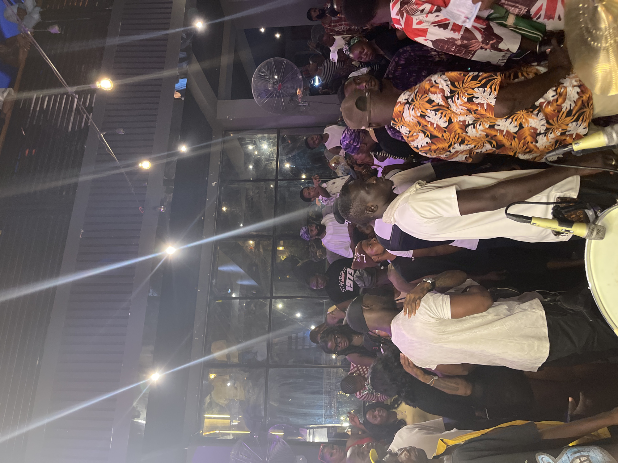 In an indoor venue with a corrugated metal ceiling and large windows, a diverse crowd of approximately 30 people is gathered. The atmosphere appears to be lively, with some individuals in the foreground wearing festive clothing such as a brightly colored floral shirt. Several people are engaged in conversation, while others are observing their surroundings. Prominent objects in the scene include a large industrial fan mounted on a wall, string lights with visible bulbs overhead, and what appears to be musical equipment, possibly part of a drum set with microphones, in the lower foreground. The lighting is dim, with spotlights creating bright streaks across the image, and the overall impression is that of a social gathering or event.