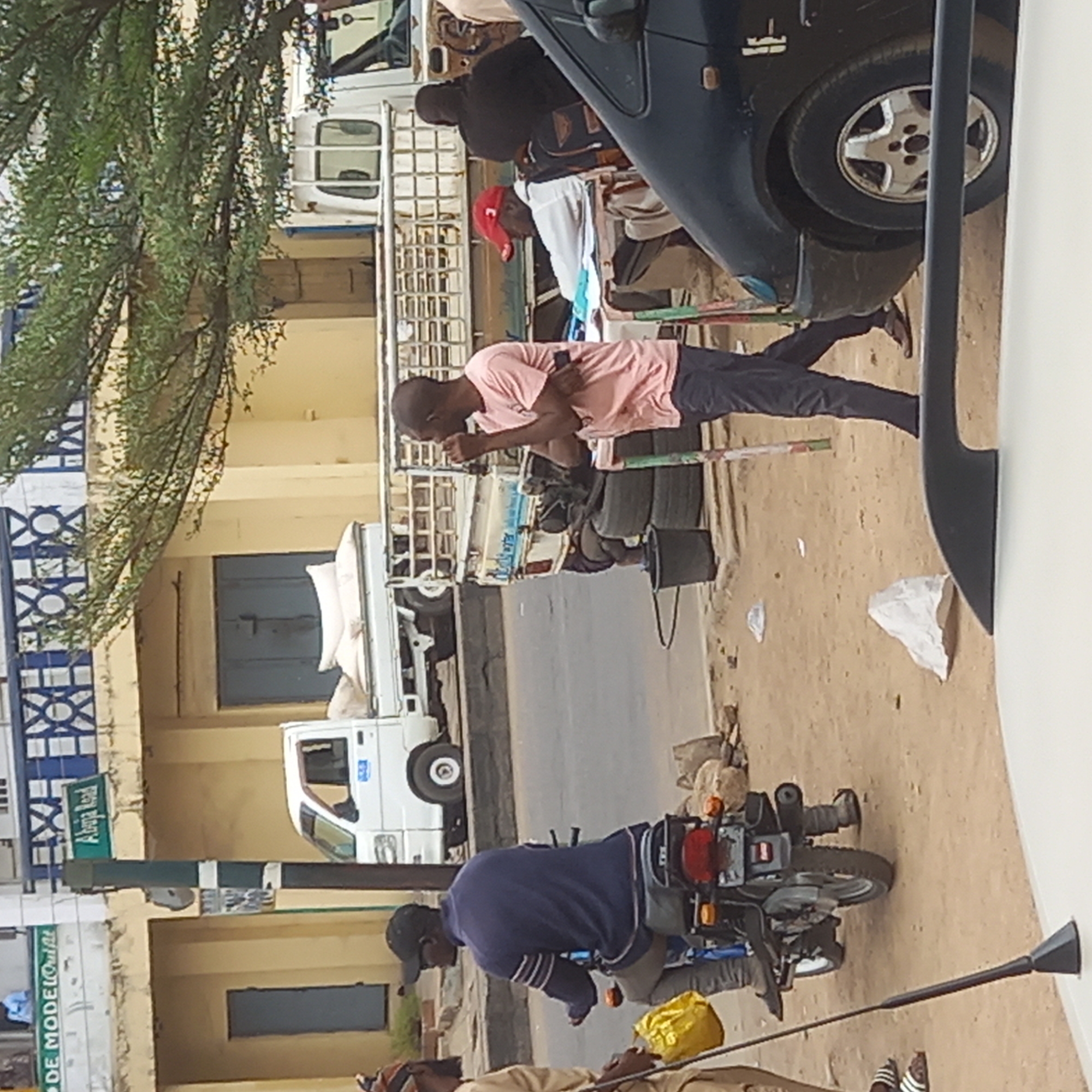 The scene depicts a street in Dadi Riba, Nigeria. A man in a pink shirt and dark pants is standing next to a vehicle and appears to be talking or eating. Several other people are present, with some visible on the back of a white truck. A man in a blue shirt is on a motorcycle in the foreground. Other vehicles are parked along the street. The road is partially paved and there is a building with blue doors and windows in the background. Signage with "Abuja Road" and "DE MODEL" is seen on a post. Trash is scattered on the ground.