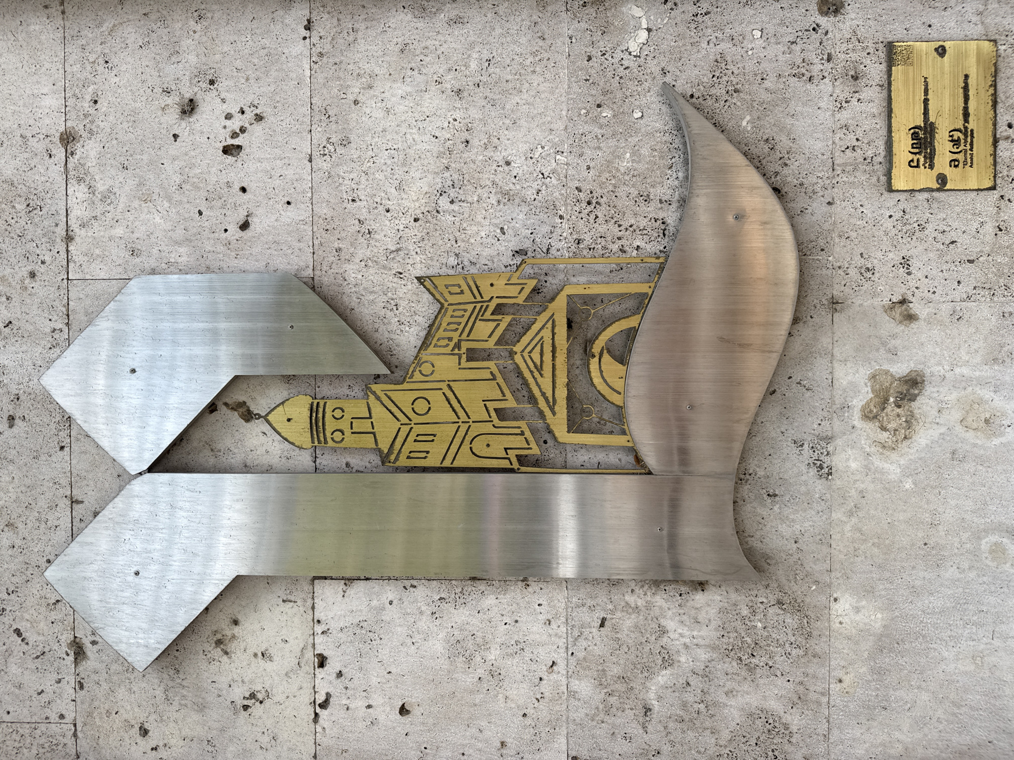 The image shows a close-up of a metallic sculpture on a textured stone wall. The sculpture is abstract, with a large "L" shape formed by brushed metal, encompassing a golden emblem. This emblem depicts a stylized building with a domed roof, resembling a church or historical structure. Below the main sculpture, a small brass plaque is attached to the wall. The plaque contains text in Armenian and English. The Armenian text is on the top section, and the English text below reads "Eternal Alphabet" and "Anahit Awakening". The stone wall appears to be an exterior surface, possibly part of a building's facade in Yerevan, Armenia, given the context of the "Eternal Alphabet" reference, which is a monument in Yerevan. The overall impression is of an artistic and informational installation on a weathered wall.