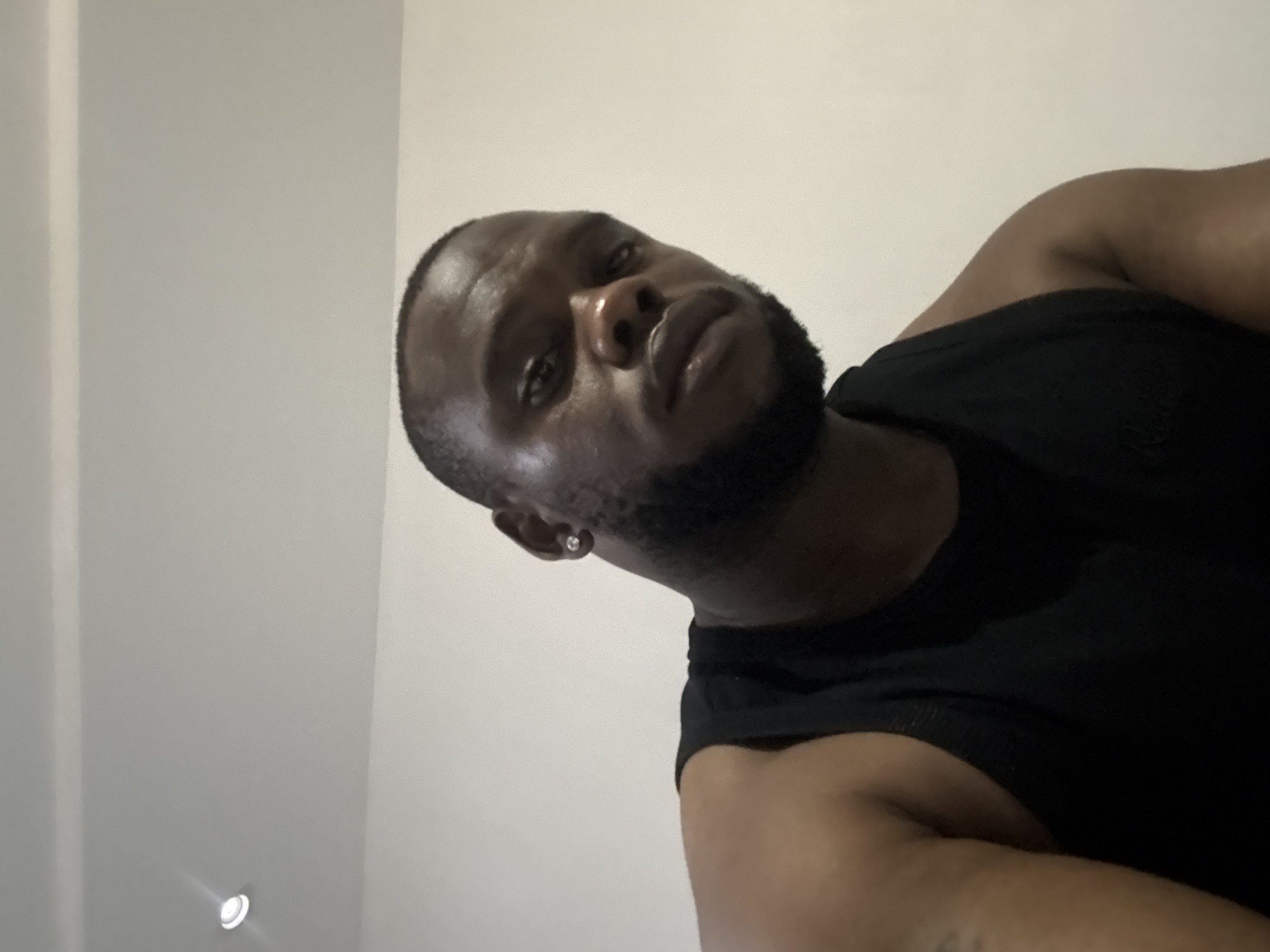 The media file displays a close-up, low-angle shot of a Black man, likely in his 20s or 30s. He is looking downwards or directly into the camera, which is positioned below him. The man has a short, dark haircut and a well-groomed dark beard. He is wearing a plain black sleeveless top or tank top. A small, sparkling stud earring is visible in his left ear. His skin appears smooth and slightly reflective, especially on his forehead and nose. A faint, unidentifiable tattoo can be seen on his right arm. His expression is neutral to slightly serious. The background is a stark, light-colored wall or ceiling, suggesting an indoor setting. In the lower left corner of the original image, a recessed light fixture with a soft glow is visible on the ceiling. There is no discernible text in the image. The image captures a personal moment, possibly a selfie.