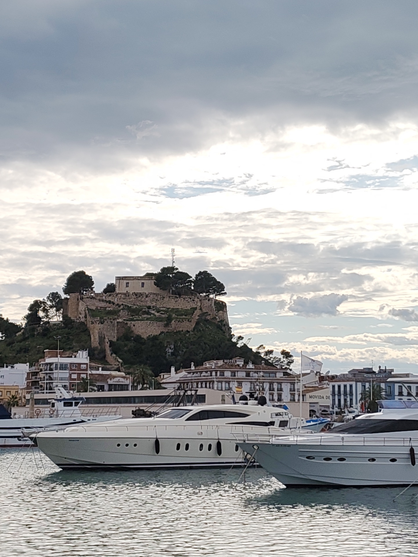 The image captures a waterfront scene in Dénia, Spain, featuring a medieval castle perched atop a hill overlooking the harbor. In the foreground, two luxury yachts are docked in the water. The city's skyline is visible, including hotels and buildings with Spanish architecture. The sky is overcast with scattered clouds.