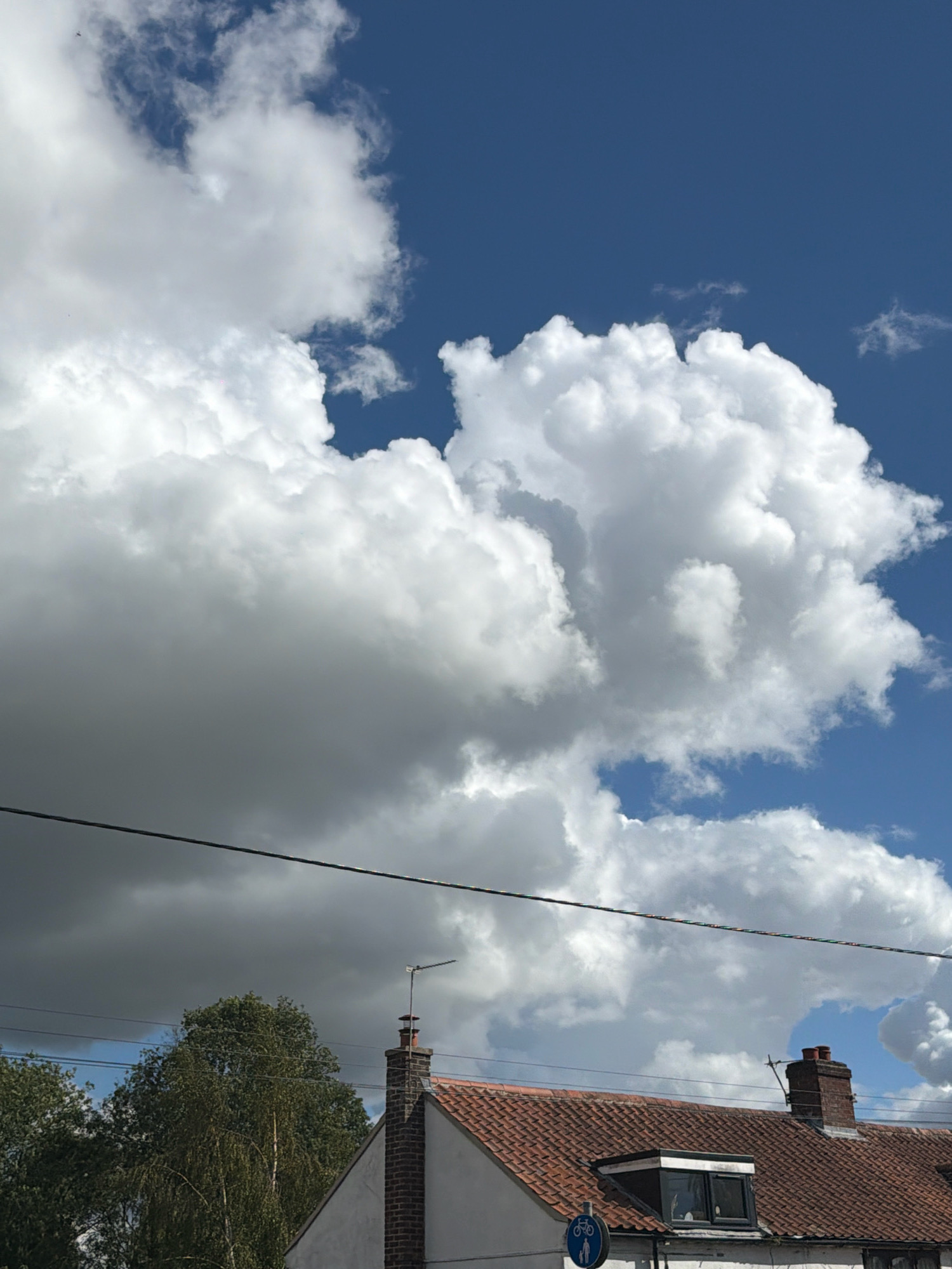 The image depicts a view of houses and trees under a dynamic sky filled with cumulus clouds and patches of blue sky. A thin power line runs horizontally across the frame, bisecting the sky and the upper portion of the scene. The roofs of two houses are visible, one with a reddish-brown tile and a chimney, and another partially visible to the left. A small dormer window with dark panes is present on the tiled roof. A traffic sign with a bicycle symbol is visible near the lower left corner of the frame. Trees with dense green foliage are present in the lower left and center of the image. The overall scene is likely captured in a residential area. The time of day appears to be daytime due to the bright sunlight illuminating the clouds and the blue sky. The presence of houses and trees suggests a rural or suburban setting, possibly within the city of South Norfolk, United Kingdom, given the context. There are no people or animals explicitly visible in the image, but distant birds can be faintly discerned in the sky.