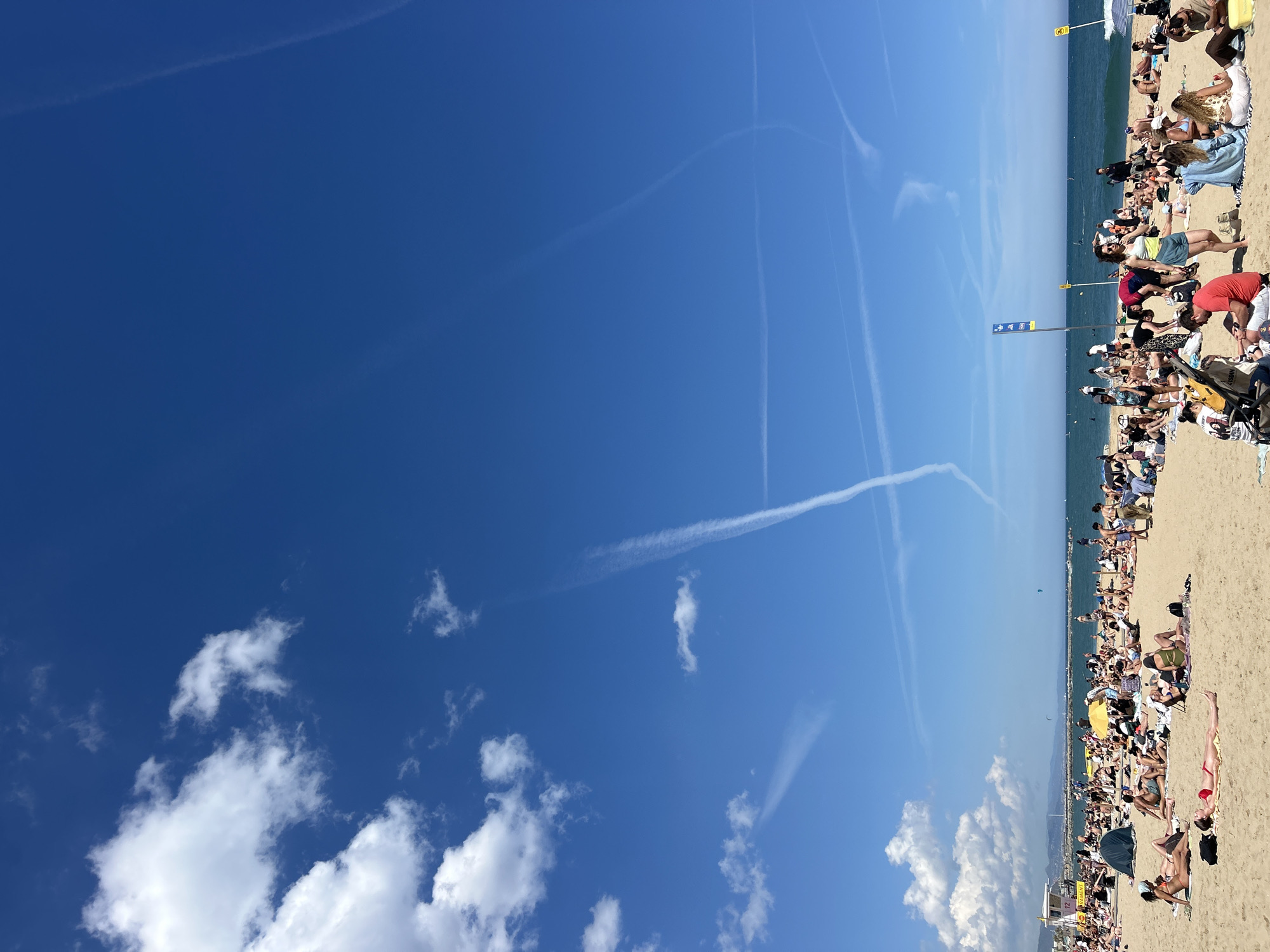 The image shows a sunny beach scene in Barcelona, Spain. The beach is crowded with people relaxing, sunbathing, and walking. The sand is light-colored, and the sea is visible in the background. The sky is blue with some clouds and contrails from airplanes. There are some poles with signs and flags along the beach, as well as some lifeguard stands.