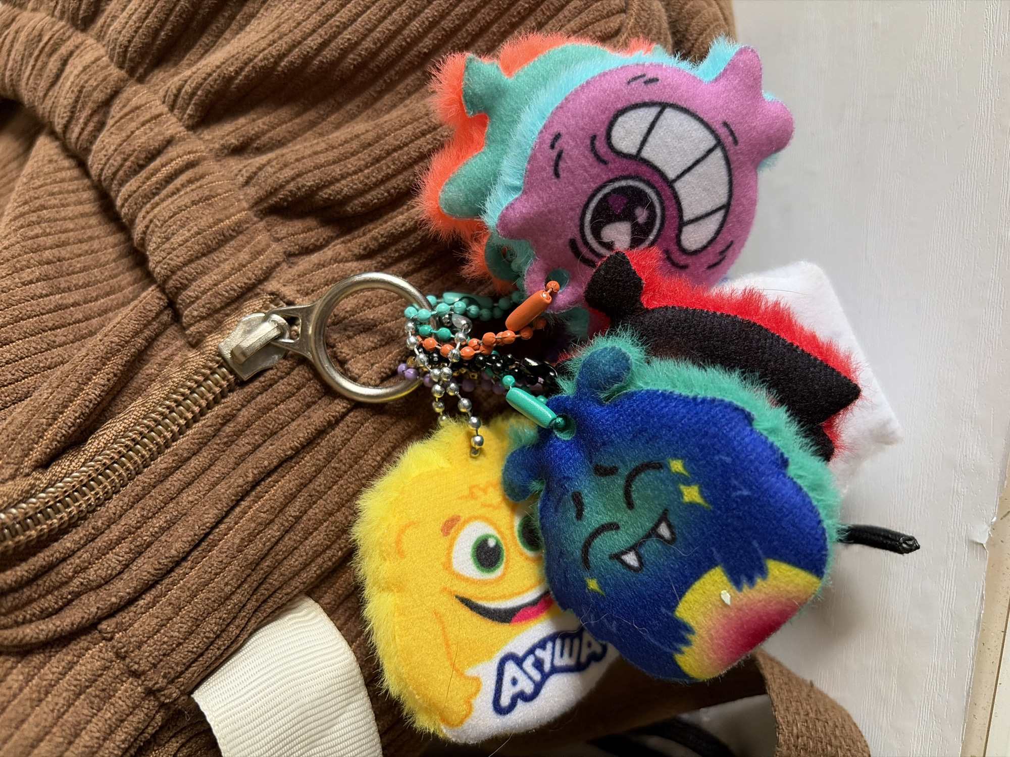 The image depicts a close-up view of a zipper pull attached to a brown corduroy garment, likely a jacket or bag. Affixed to the zipper pull are multiple plush keychain charms. These charms are colorful, fuzzy, and depict cartoon-like monster faces. One charm features a yellow character with a wide smile and the word "Агуша" (Agusha), which is a Russian brand of baby food. Another charm is blue with small stars. The remaining charm is a layered creation of pink, teal, and orange on top of a base of black, red, and white. The composition suggests that the objects are lying on a flat surface, next to a white wall. This location in Izhevsk, Russia can not be verified by the visual data.