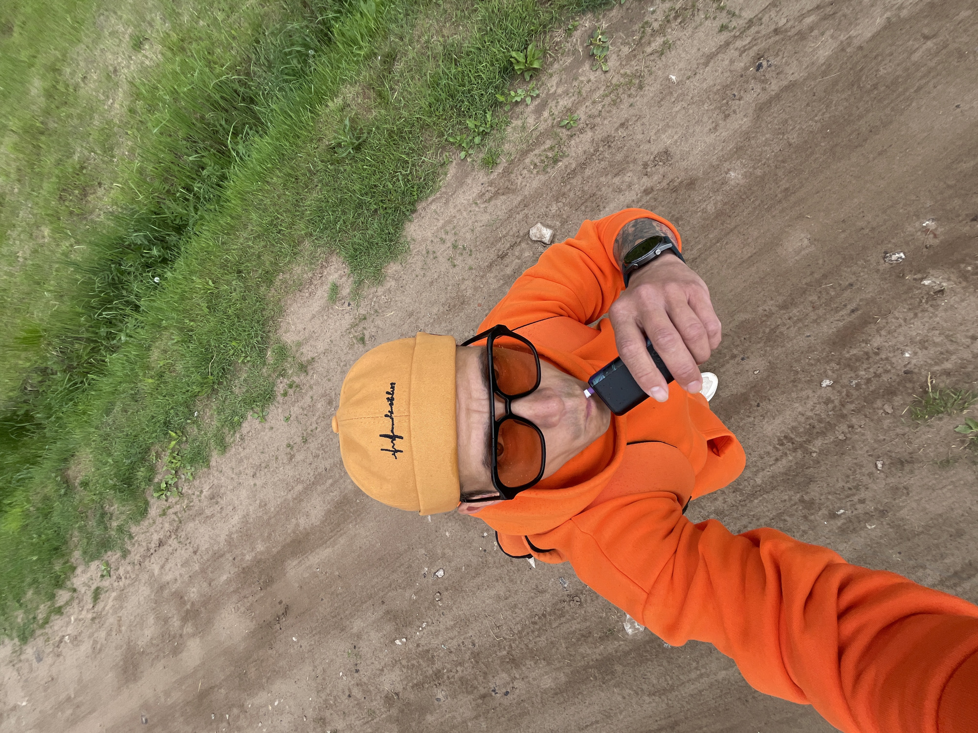 An overhead shot captures a person standing outdoors on a dirt path next to a grassy area. The subject is a male, seen from the chest up, looking slightly upwards towards the camera. He is wearing a bright orange hoodie and a mustard yellow or orange beanie. The beanie has black embroidered text on it, appearing to be "life of a boss" or a similar stylized script. He wears dark sunglasses with orange-tinted lenses. On his left wrist, a black smartwatch is visible. In his right hand, he holds a small, black electronic device, possibly a vape or a portable charger. The ground beneath him is a mix of dry, light brown dirt with small stones, and vibrant green grass lines the left side of the frame, suggesting a natural or semi-natural outdoor environment. The lighting indicates it is daytime. The scene could be a rural path or an unpaved area in a park, located in Chaikovsky, Russia.
