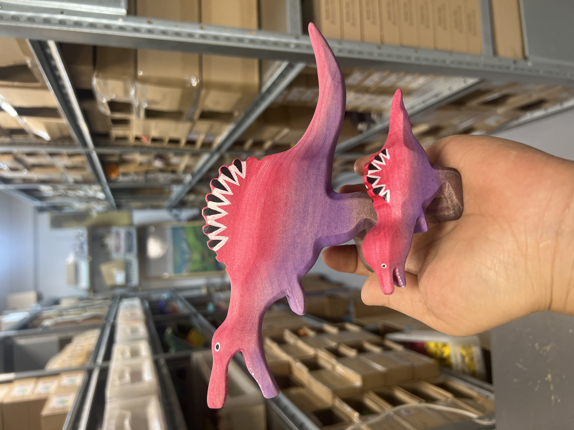 A close-up shot shows a hand holding two wooden toy dinosaurs, with a blurred background of shelves filled with boxes. The larger dinosaur, in the foreground, is pink and purple, with a white and black-striped sail on its back. The smaller dinosaur, held lower down, is also pink and purple with a similar sail. The hand is slightly cupped, supporting the toys. The background features rows of metal shelves stacked with cardboard boxes of various sizes, indicating a warehouse or storage facility. Some boxes appear to be wrapped in plastic.