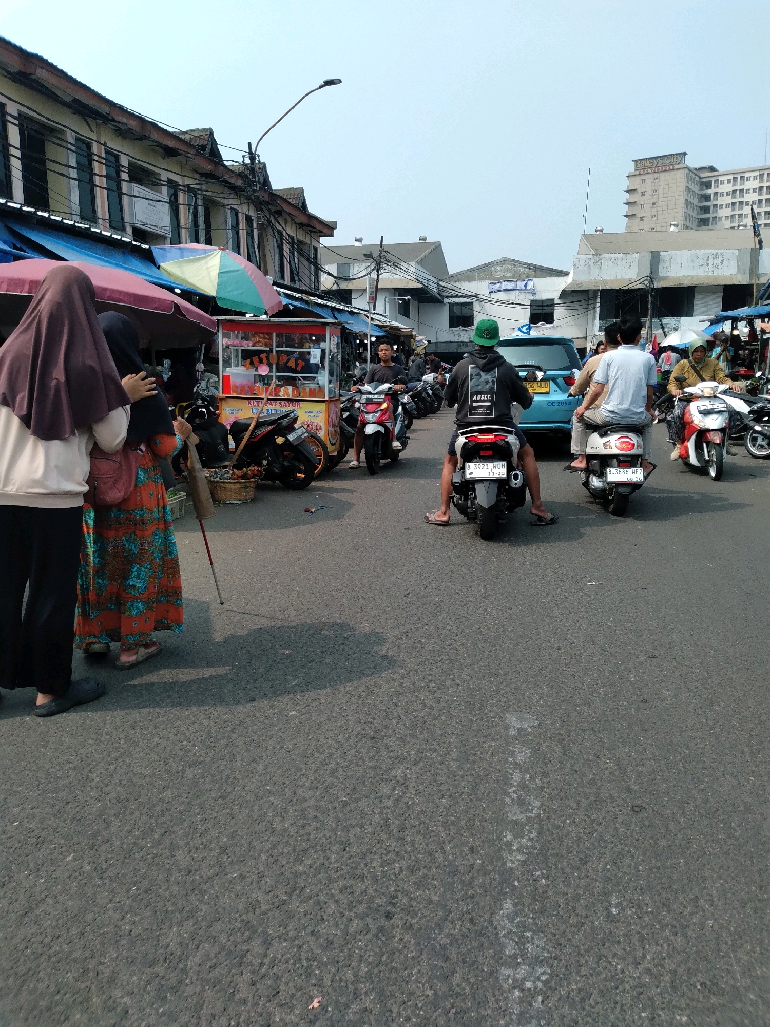 This is a street-level, medium-distance, eye-level shot of a busy market scene in South Tangerang, Indonesia, on a bright, clear day. The foreground is dominated by an asphalt road, with a partial shadow of a person on the right. In the mid-ground, several individuals are on motorcycles. On the left, two women, one wearing a hijab and the other holding a walking stick, stand near a food stall with a colorful umbrella. Behind them, a cart sells food with clear signage, and a motorcycle is parked. Further back, a blue car is present, and more people are on motorcycles, including a man in a green cap. The background features a row of aging buildings with awnings and utility wires, and a taller modern building labeled "Bailey's City" in the distance. The atmosphere suggests a typical bustling market environment with pedestrian and vehicular traffic.