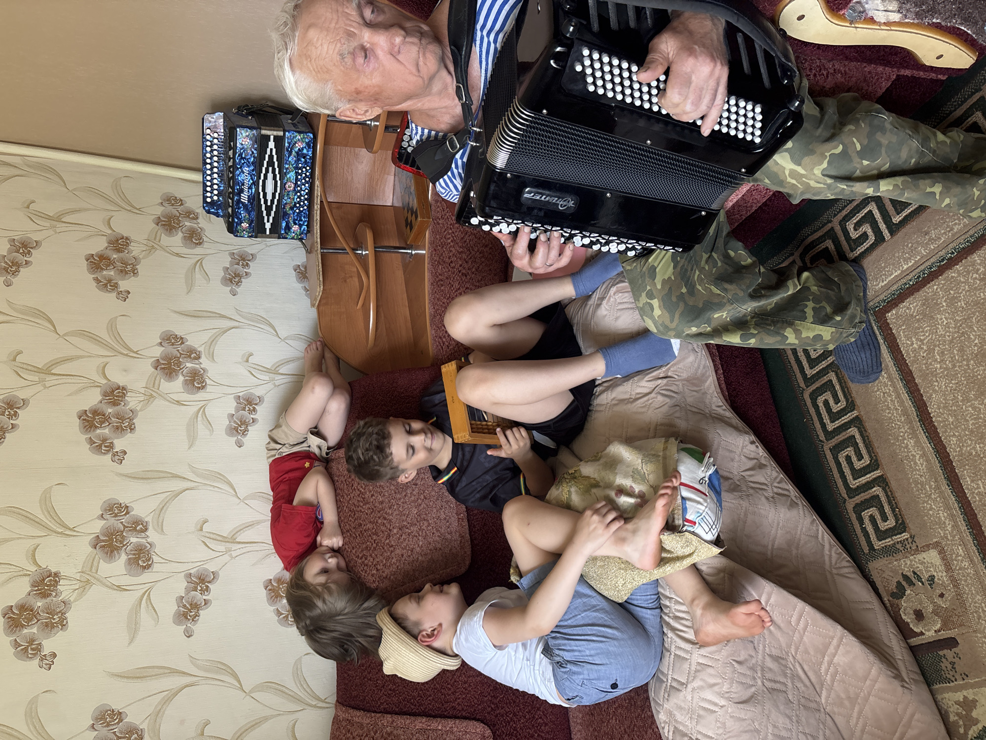 The image depicts an elderly man playing a black accordion while seated on a couch with at least three children lying around him. He is wearing a striped shirt and camouflage pants. The children are dressed in casual clothing. A smaller accordion sits on a wooden stand near the wall. The environment is a room with patterned wallpaper. The location is Маркидоновка, Russia.