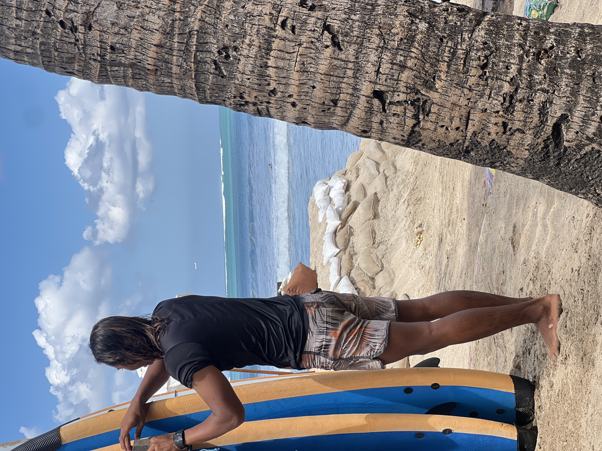 A person is standing on a sandy beach next to a blue and yellow surfboard, facing the ocean. The person is wearing a black t-shirt and patterned shorts. The ocean has waves, and there are sandbags piled on the beach in the background. A palm tree trunk is visible in the foreground on the right side of the image. The sky is blue with white clouds, and a small airplane is visible in the distance. The image appears to be taken in Kuta, Indonesia, a popular tourist destination known for its beaches and surfing.
