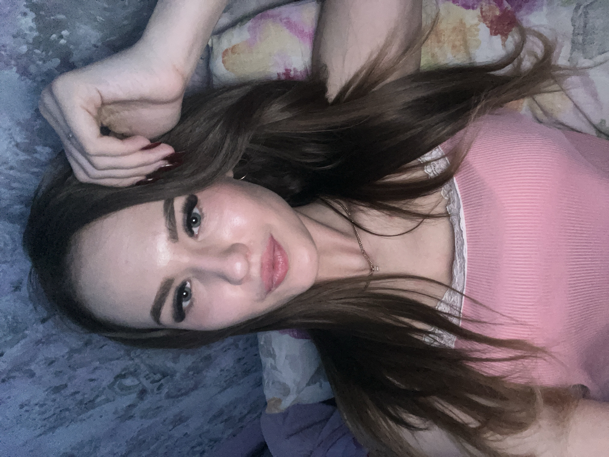 A woman with long brown hair is lying on a patterned bed. She is wearing a pink top with white lace trim. Her left hand is raised near her head. The bedding has a floral pattern in cream, pink, and purple tones, while the sheet underneath is light blue with darker blue patterns. The location is unknown; there is no evidence to suggest this image was taken in Noviy, Russia.