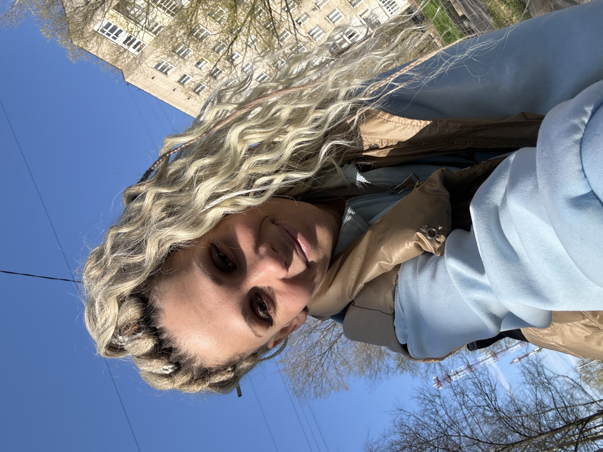 A selfie perspective shot shows a woman with long blonde hair styled in braids and waves. She is wearing a light blue hoodie and a tan puffy vest or jacket. The background shows a clear blue sky, bare tree branches, and a multi-story building with many windows. The presence of a crane or construction equipment is also evident among the tree branches. Based on the description, the photograph was taken in Rusanovo, Russia.