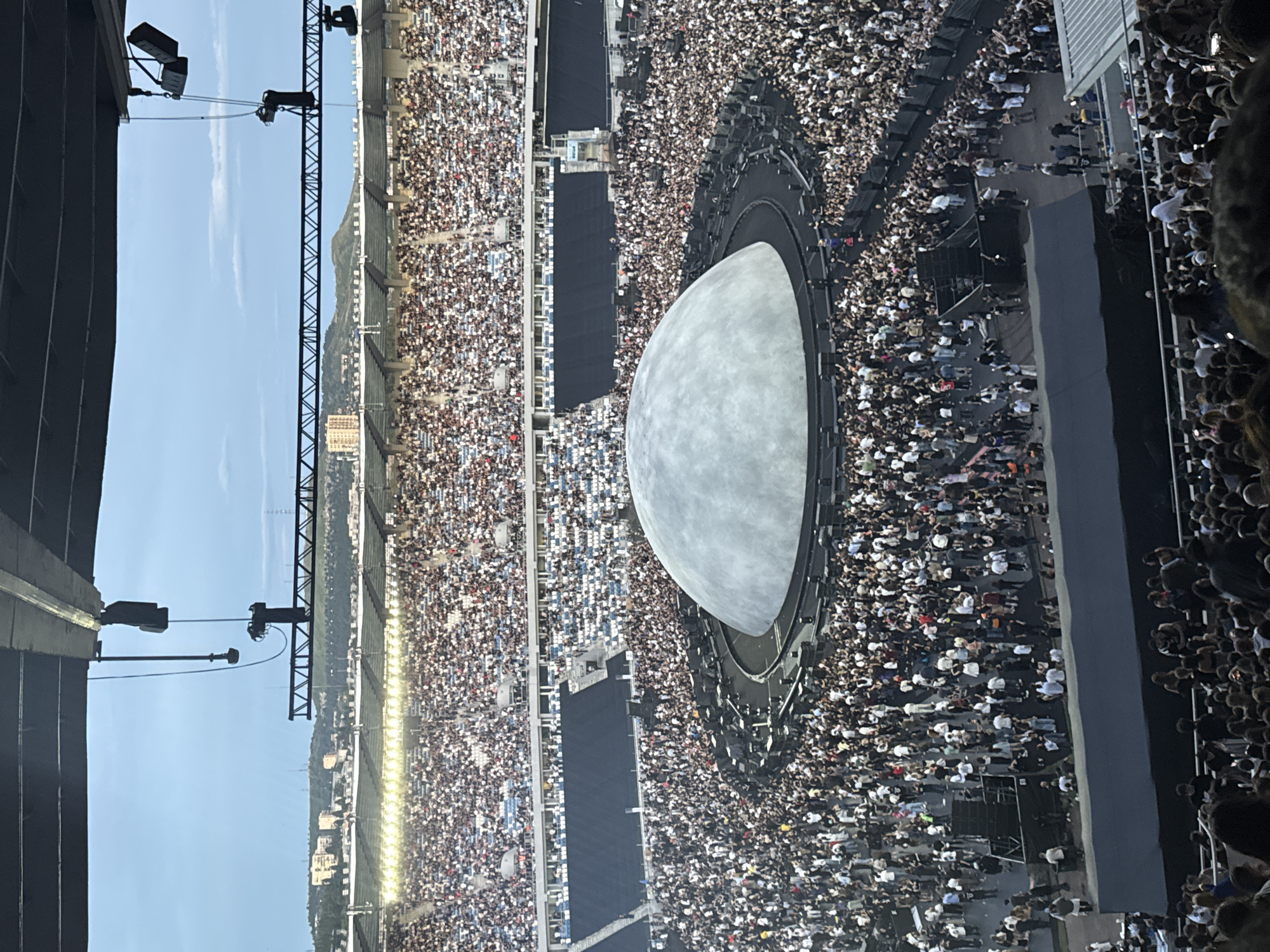 This image depicts a large outdoor concert venue, likely a stadium, filled with a substantial crowd of spectators. In the center of the field, a prominent, large, dome-shaped stage structure is visible, surrounded by a circular platform and various technical equipment. The stands are packed with people, suggesting a high-attendance event. The background shows a cityscape with buildings and hills under a cloudy sky, consistent with the location of Tbilisi, Georgia, known for its hilly terrain and architectural style. The overall scene conveys the atmosphere of a major live musical performance.