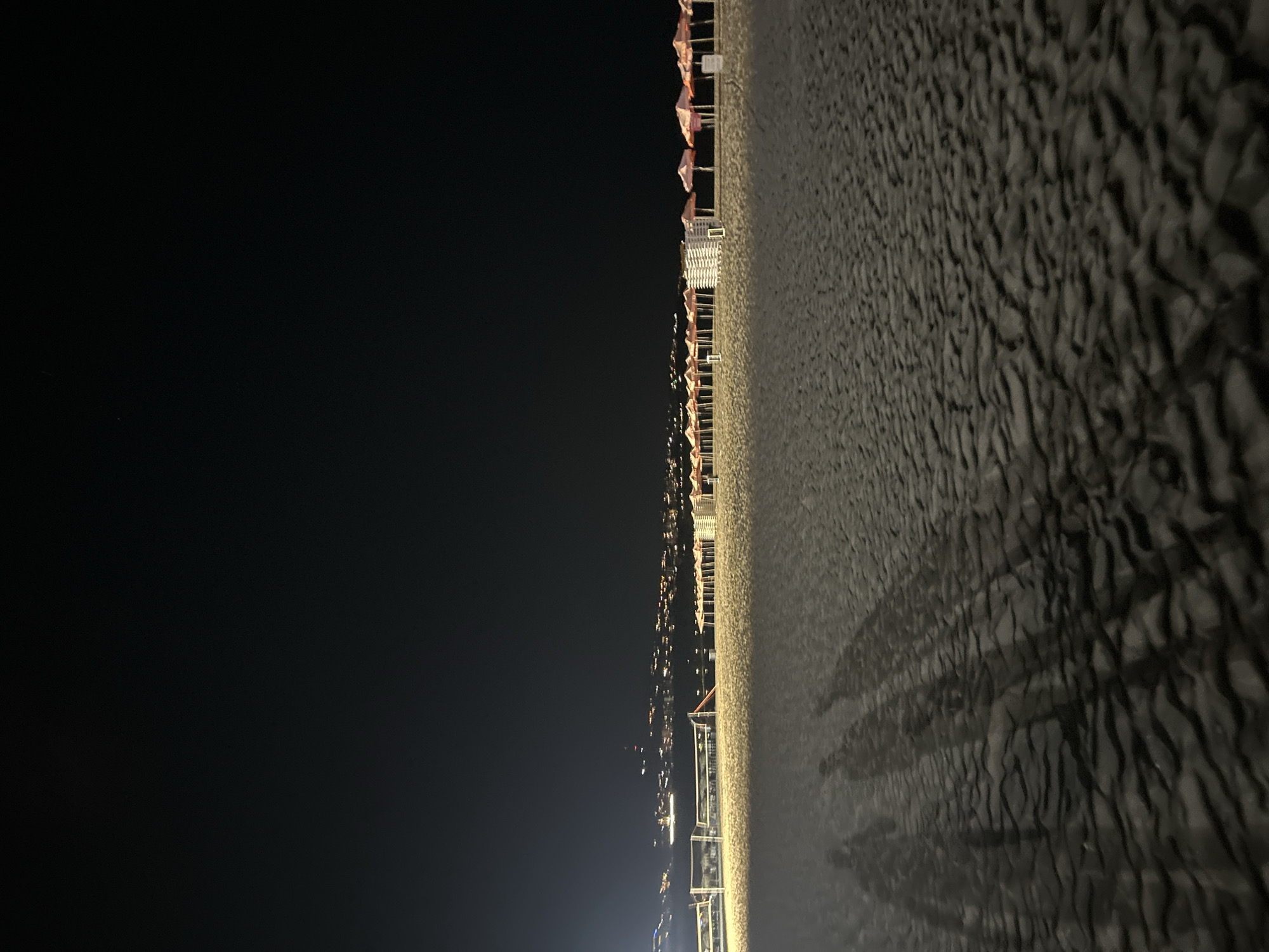 The image captures a night scene on a sandy beach in Varna, Bulgaria. The foreground is dominated by sand dunes, with the elongated shadows of three individuals cast upon them by an unseen light source. In the middle ground, a row of beach umbrellas, predominantly pink, are neatly arranged, interspersed with stacks of what appear to be lounge chairs. Further in the distance, a line of lights illuminates a cityscape or waterfront area. A volleyball net is visible on the left side of the frame, suggesting recreational facilities. The overall ambiance is one of a quiet, nocturnal beach setting.