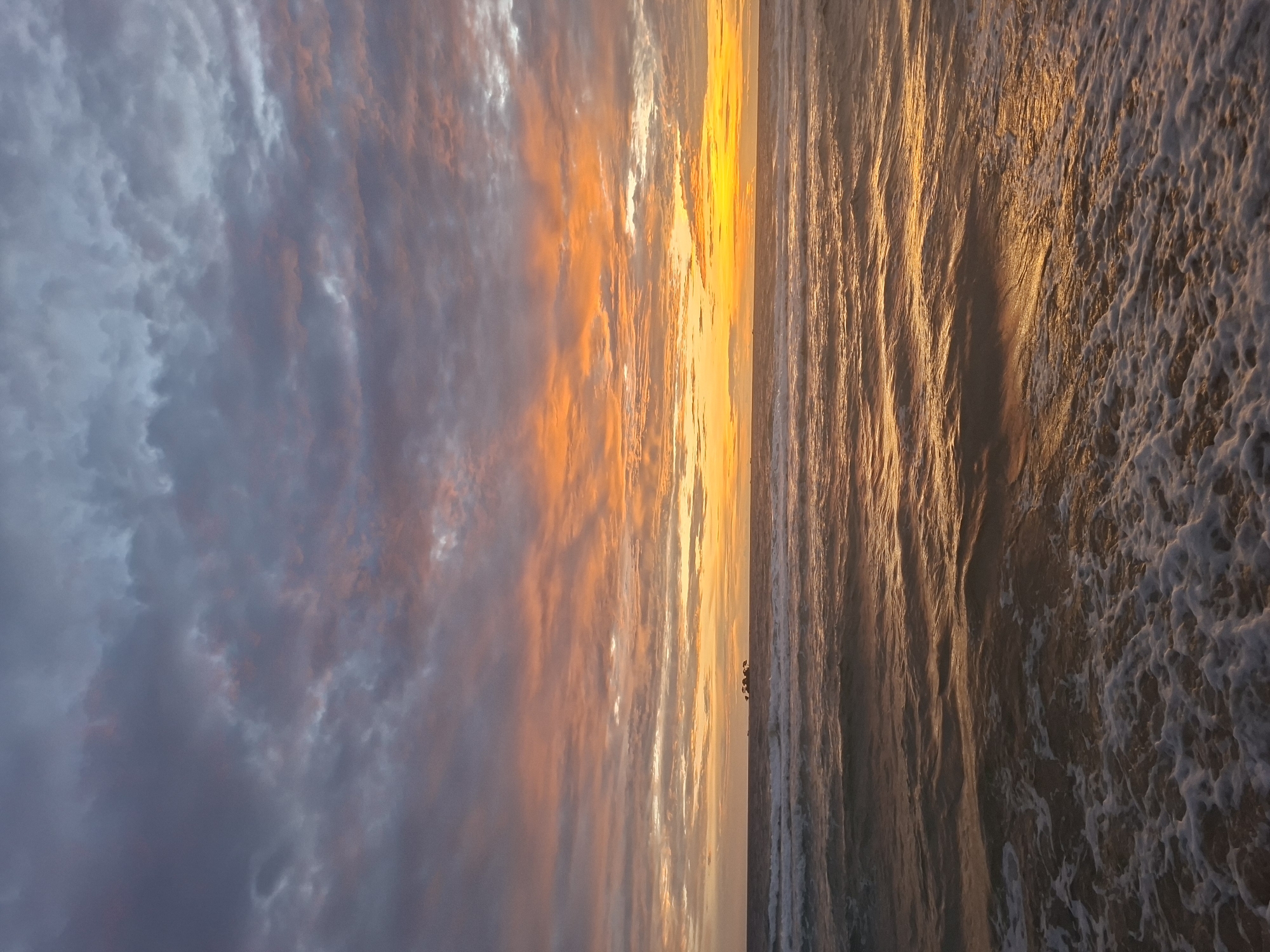 The image depicts a coastal scene in Phú Quốc, Vietnam, during either sunrise or sunset. The sky is filled with dramatic clouds, transitioning from a deep gray and purple at the top to vibrant shades of orange and yellow near the horizon. The sun's rays are casting a warm, golden glow across the ocean, creating shimmering patterns on the water's surface. Gentle waves are rolling towards the shore, with foam accumulating in the foreground. In the distance, a small cluster of dark, rocky formations topped with sparse vegetation is visible on the sea. The overall atmosphere is serene and picturesque, characteristic of a tropical coastal landscape. There are no people or specific urban structures visible in this particular frame.