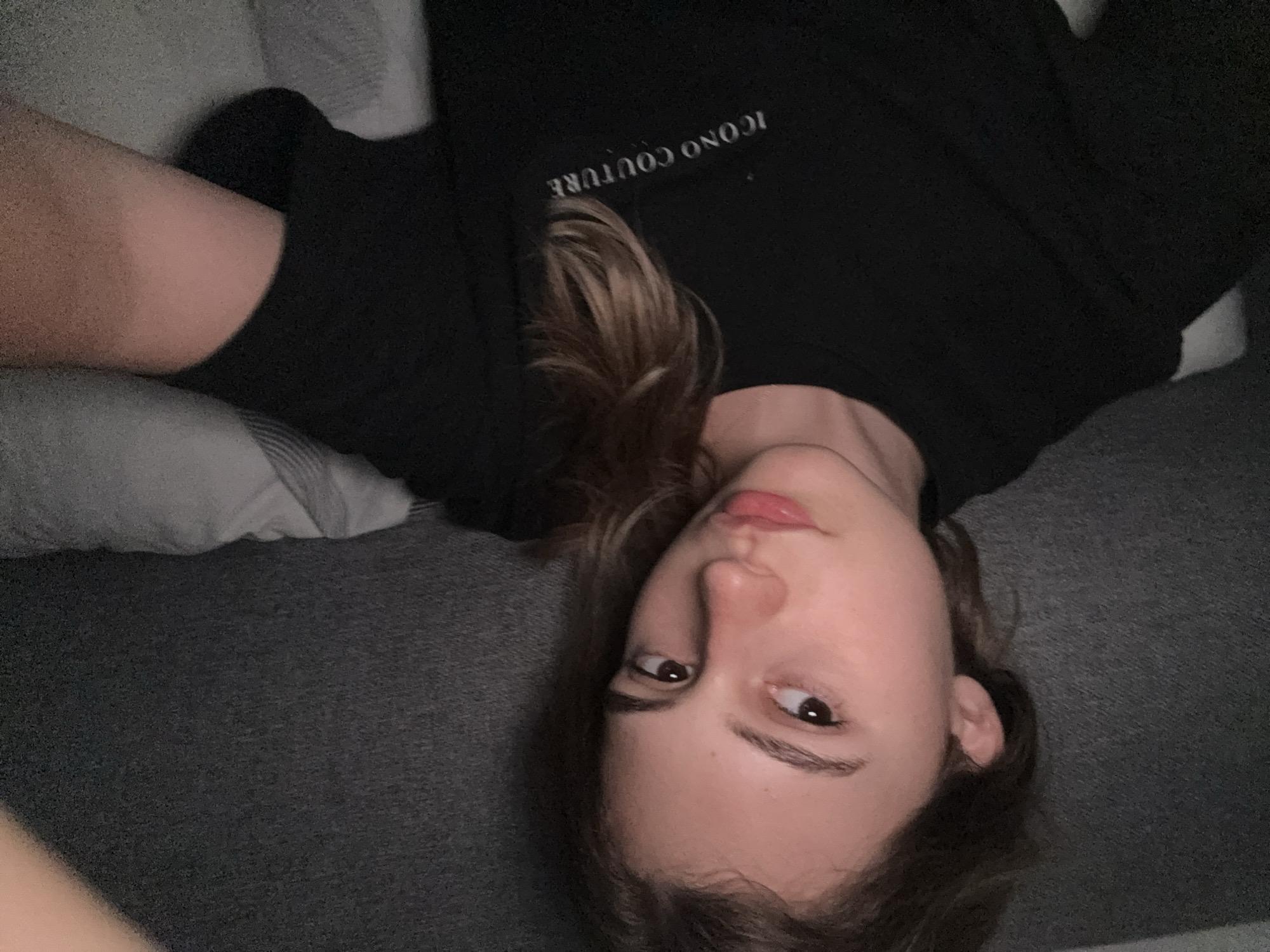 A young Caucasian female is pictured from the shoulders up, positioned slightly to the left of the frame. She has light brown hair that falls loosely around her shoulders and face, with some strands framing her features. Her skin appears fair, and she has light brown eyes and full lips. She is wearing a black t-shirt with the words "ICONO COUTURE" printed in a lighter color across the chest. The background consists of a dark, textured wall and what appears to be a grey, striped pillow or bedding behind her. The lighting is dim, suggesting an indoor setting, possibly a bedroom. There is no indication of the location being in the city of Forbach, Germany, or any specific urban environment.