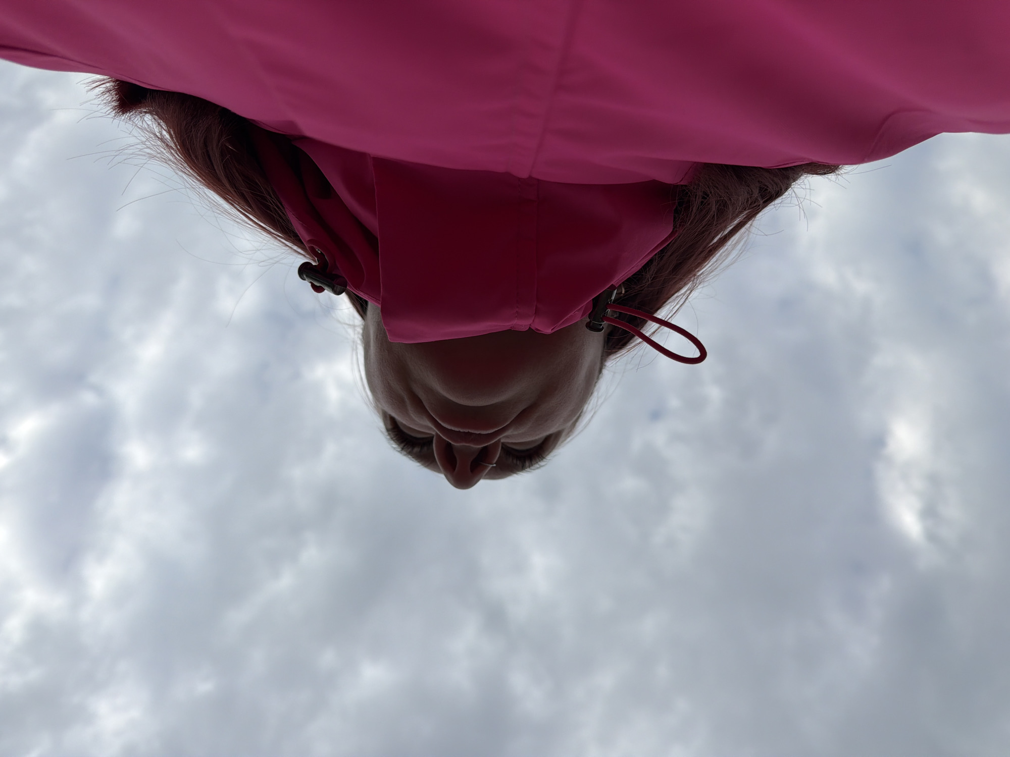 The image is an upward-facing perspective shot of a person standing outside. The person's face, from the nose and eyes up, is visible against a backdrop of a cloudy sky. The person is wearing a pink jacket with a hood. The location is unknown, and there is no indication of it being Chaikovsky, Russia.