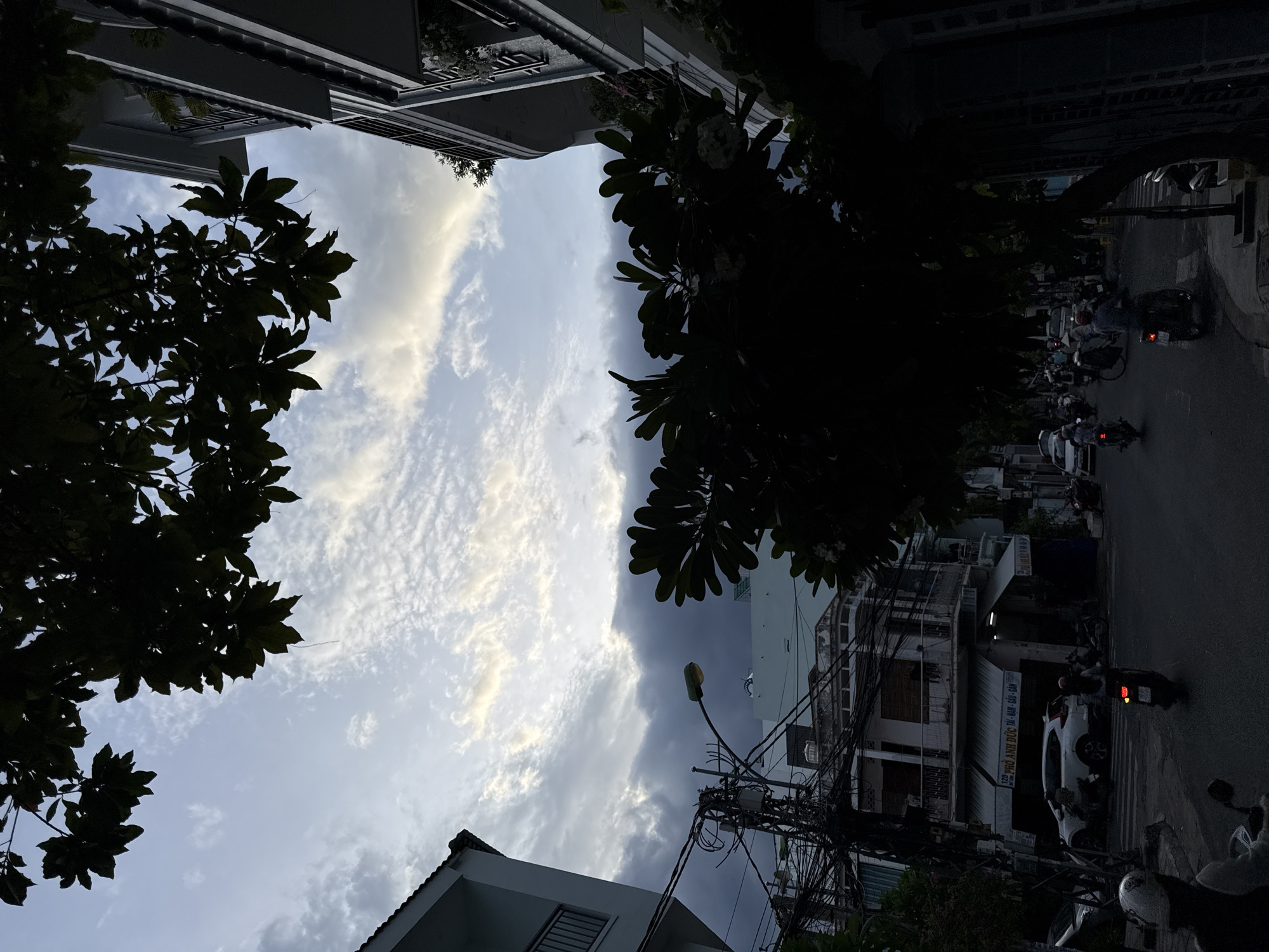 The image depicts a street scene in Nha Trang, Vietnam, at dusk or early evening. The sky is filled with dramatic clouds, with patches of sunlight breaking through, creating a luminous effect. In the foreground, silhouetted against the sky, are the dark foliage of trees and branches.
On the left side of the image, a white building with balconies is visible. Adjacent to it, a complex network of electrical wires and poles dominates the mid-ground, characteristic of many urban environments in Southeast Asia. Below the wires, a two-story building with shops on the ground floor can be seen, one of which has signage in Vietnamese. A white SUV is parked in front of one of the shops, and several motorcycles are on the street.
The street itself is paved and appears to be a busy thoroughfare. Various people are riding motorcycles, some with helmets. The atmosphere is dimly lit, suggesting either the approaching night or overcast conditions. The right side of the image shows another building with balconies, also partially silhouetted by the trees. The overall impression is one of a lively urban street experiencing a transition in lighting.