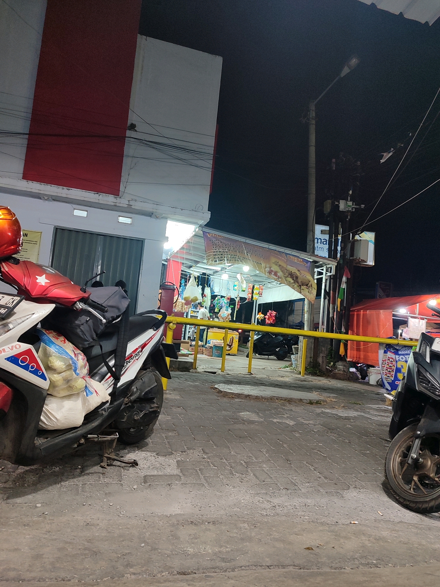 The image depicts a street scene at night in Garut, Indonesia. A red and white building is visible on the left, with a prominent red stripe. In the foreground, a white and red scooter is parked, carrying a large bag of items. Another black scooter is partially visible on the right. Various stalls and structures are set up along the street, illuminated by artificial lights. A bright orange tent is visible further down the street. The ground is paved with cobblestones. Electrical wires are strung across the scene, and a street lamp illuminates the area. The overall impression is of a busy, albeit nocturnal, commercial area.