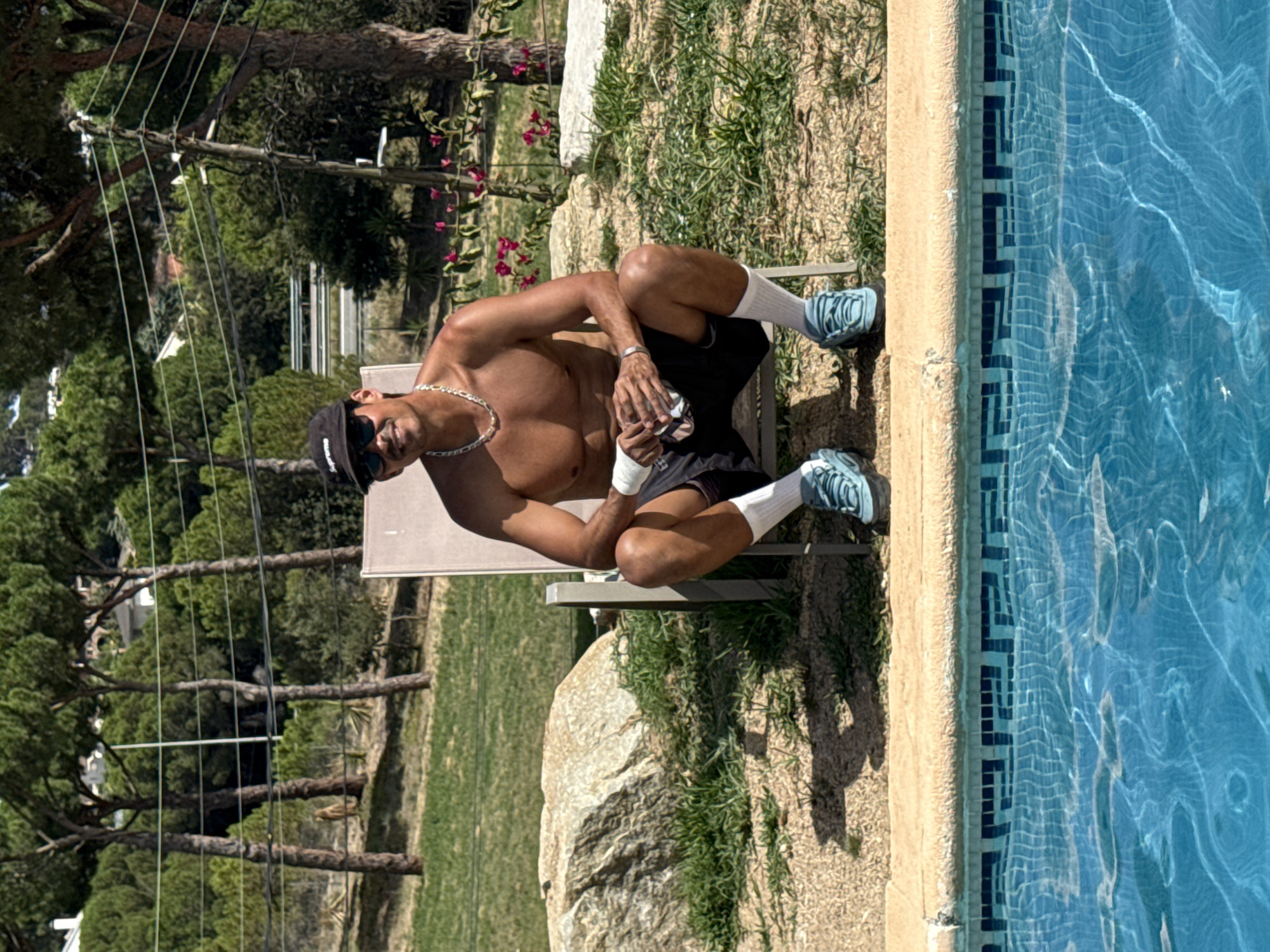 A man is sitting on a lounge chair by a swimming pool. He is shirtless, wearing black shorts, white socks, and blue sneakers. He has a black baseball cap and sunglasses on and is wearing a silver chain. The man is holding a clear cup. The lounge chair is beige and is on a paved area next to the pool. Behind the man is a grassy area with trees and rocks. The swimming pool has blue water with white lines on the bottom and a tiled edge. The scene appears to be a backyard or resort in Sant Vicenç de Montalt, Spain.