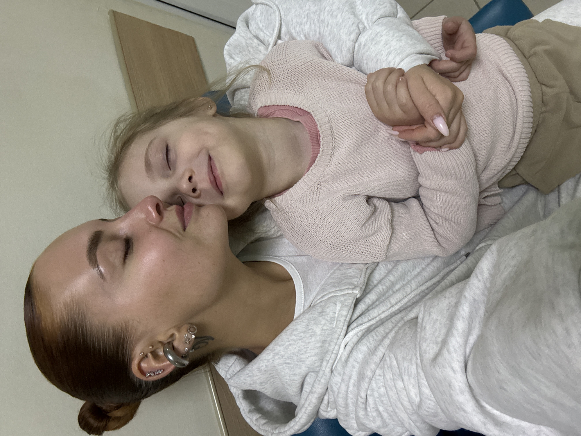 An adult and a young child share a tender, intimate moment, lying down close together, their faces almost touching. Both have their eyes closed, suggesting a moment of peace, rest, or deep affection. The adult, positioned in the foreground on the left, has light brown hair pulled back in a bun and numerous intricate ear piercings, including a large silver conch ring and several helix and lobe piercings. She wears a light grey hooded sweatshirt.
The child, with blonde hair, is nestled against the adult, wearing a light pink knitted sweater over a darker pink undershirt and light brown pants. A faint, peaceful smile plays on her lips. Their hands are clasped together between them, with the adult displaying long, light pink manicured nails. The setting appears to be indoors, likely a home, with a light-colored wall and what looks like a wooden panel in the background. A blue surface, possibly a pillow or part of a piece of furniture, is visible underneath them. The overall scene conveys warmth, comfort, and a strong emotional bond. There is no visible text in the image. The location context is Chaikovsky, Russia.
