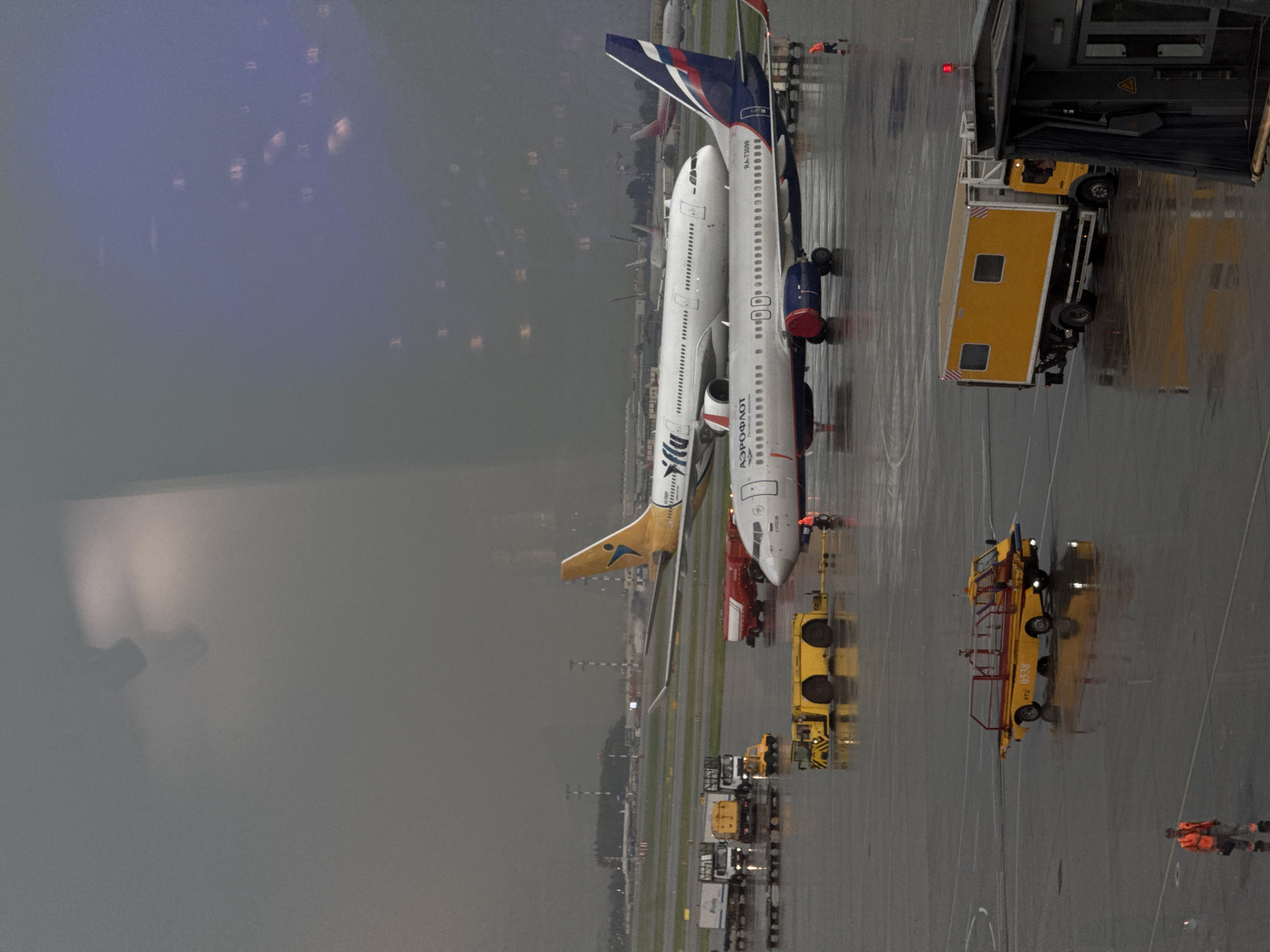The image depicts an airport tarmac in Khimki, Russia, on a rainy day. Several aircraft are visible, including a white passenger plane with "S7 Airlines" written on its fuselage, and another white plane with a blue and red tail fin belonging to Aeroflot. Various airport vehicles are also present, such as baggage tugs, fuel trucks, and a yellow vehicle that appears to be a tow tractor. The ground is wet and reflective due to the rain, and there are puddles on the tarmac. In the background, airport infrastructure, including control towers and other aircraft, can be seen through a hazy atmosphere. Two individuals in high-visibility orange vests are present on the tarmac, one standing near an Aeroflot plane and the other walking in the foreground. The overall scene conveys a busy operational environment at an airport despite the inclement weather.