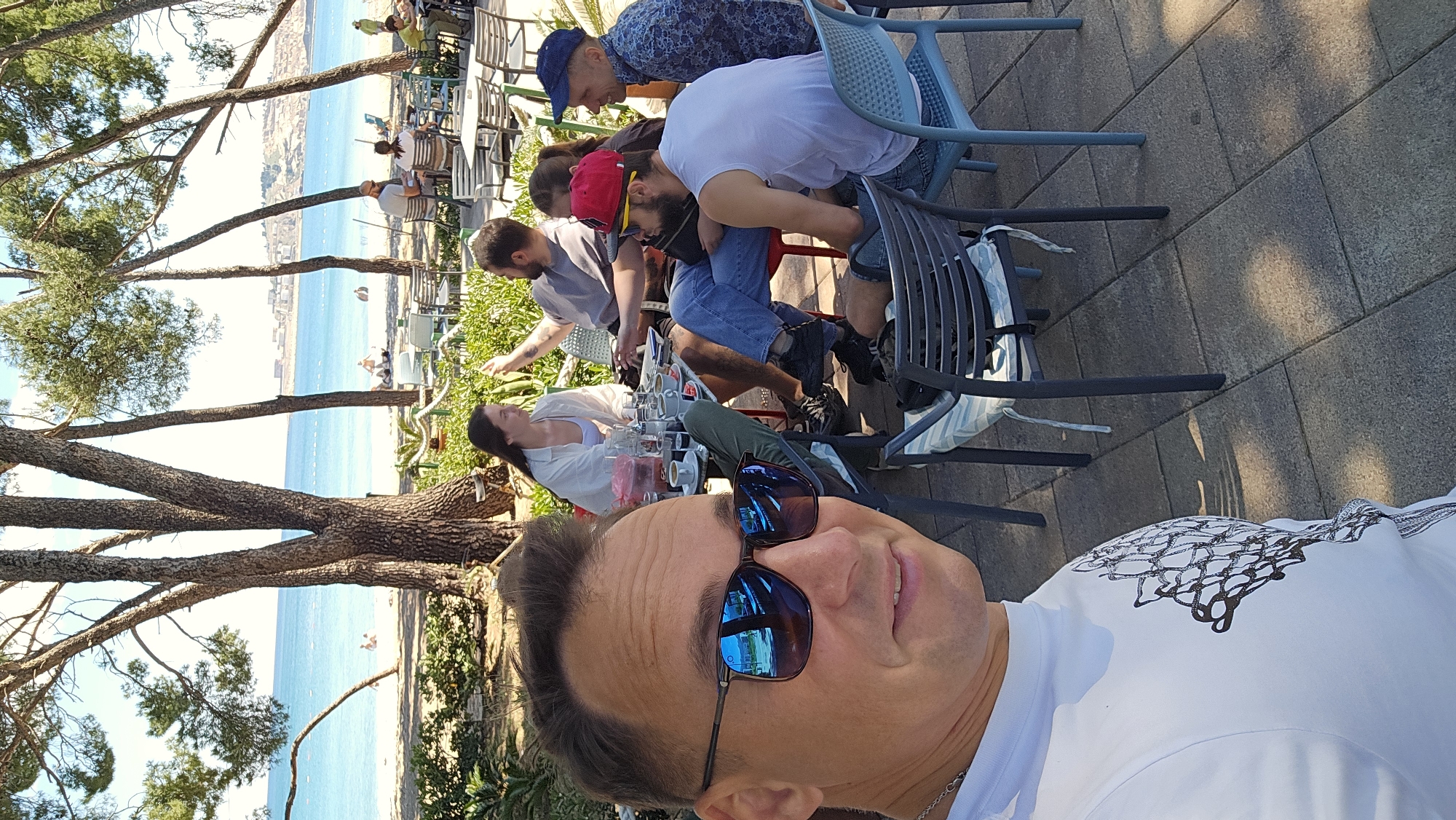 The image is a selfie of a Caucasian male wearing sunglasses and a white t-shirt with a design on it. He is smiling slightly. Behind him, there's a group of people sitting at tables in what appears to be an outdoor café or restaurant setting. One person is wearing a red hat, and another is wearing a blue patterned hat.
In the background, there's a view of a beach and the sea. There are trees and buildings visible along the coastline. Given the mention of Bar, Montenegro, in the prompt, it's presumed that this scene is located in Bar. The people in the background appear to be engaged in conversations or observing the surrounding scenery. There are beverages on the table. The scene is illuminated by sunlight.