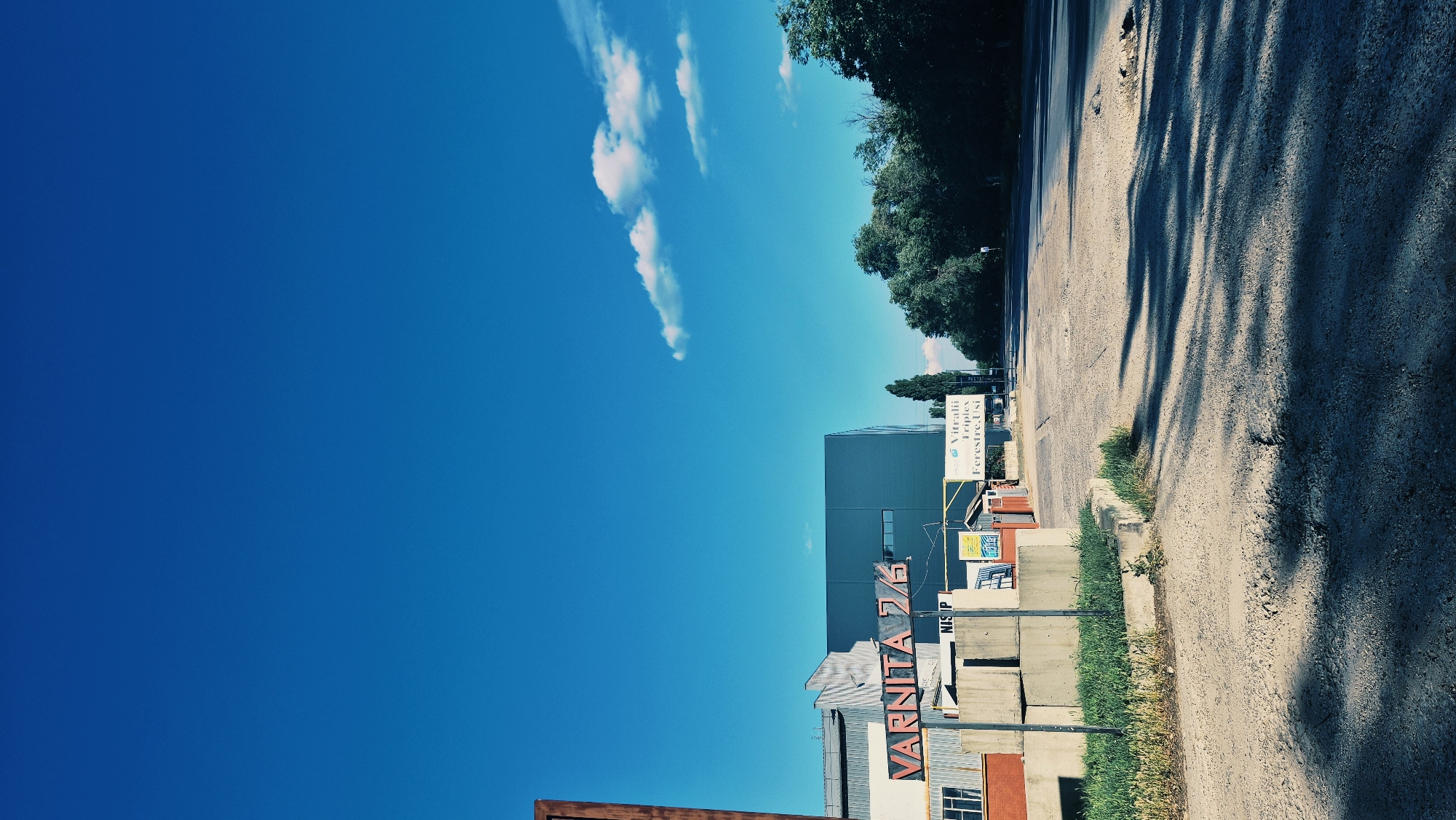 This is a vertical, outdoor, daytime photograph taken in Chișinău, Moldova. The scene depicts a roadside with a building, vegetation, and a bright blue sky with sparse white clouds. The image is oriented with the sky occupying the left two-thirds and the ground and structures on the right.
In the foreground on the right, a gravel road runs diagonally away from the viewer, casting long, sharp shadows from unseen trees. A concrete curb separates the road from a strip of green grass. Behind the grass, there are stacks of concrete blocks and some building materials.
A prominent, dark blue, modern building is visible to the left of the road, featuring several horizontal windows. To the right of this building, several signs are displayed. One sign, with a white background and blue lettering, reads "Vitrali Triplo Ferestre Uși". Another sign, with red and white lettering, reads "VARNITA". A smaller sign with blue text on a yellow background is also visible.
The time of day appears to be late morning or early afternoon, judging by the length and direction of the shadows. The weather is sunny and clear, with a vibrant blue sky. The overall mood is one of a commercial or industrial area on a pleasant day. There are no people visible in the image.