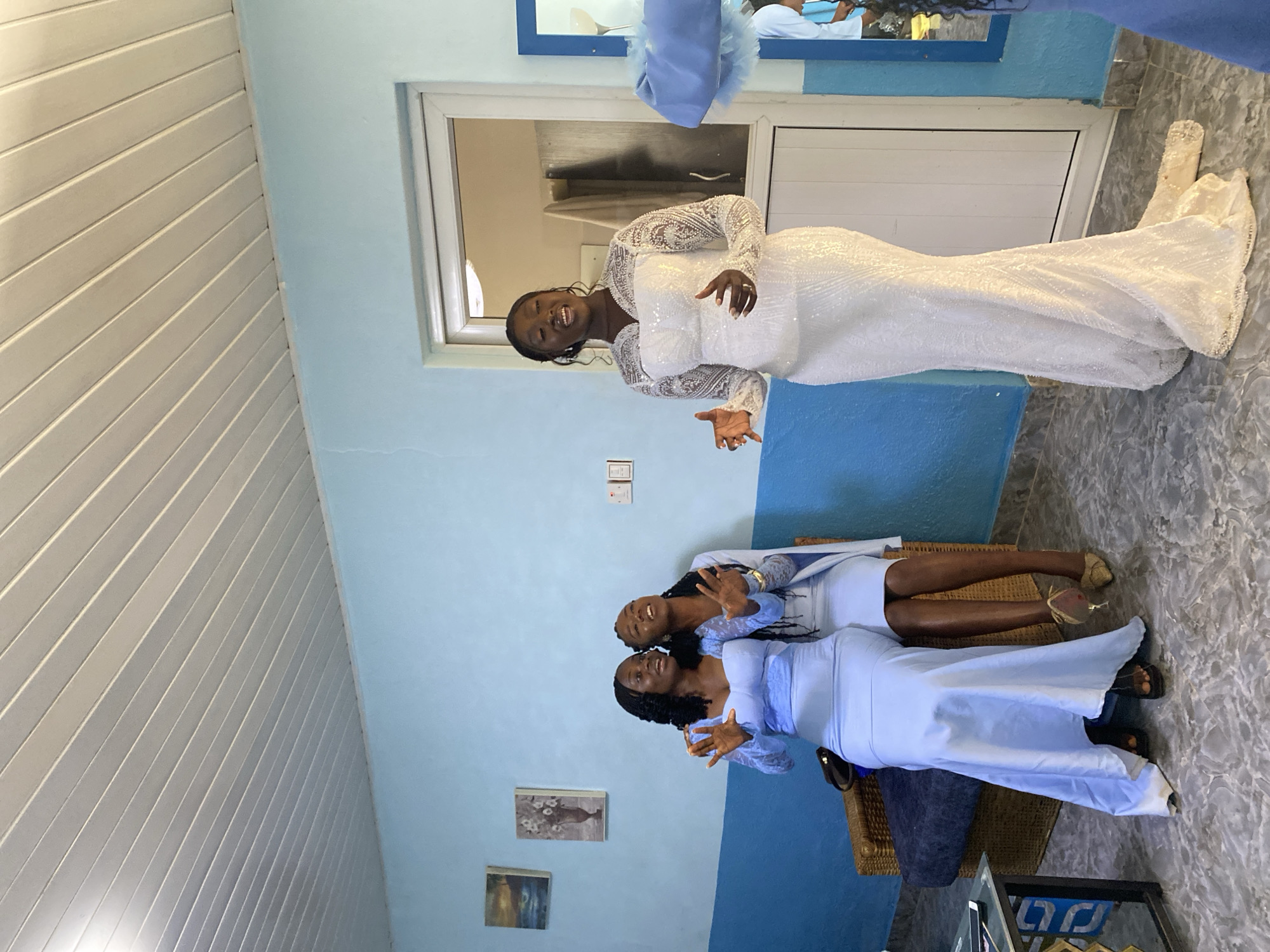 Three women are posing indoors. One woman is standing in a white, long-sleeved, floor-length dress. Two women are seated side-by-side in pale blue dresses. The walls are light blue with a dark blue stripe near the floor. There are pictures on the wall. The floor is tiled in a gray, speckled pattern. There is a white paneled ceiling. There is a white door with a white frame in the background. There is a blue frame at the top of the picture. The location may be Barakin Angundi, Nigeria.
