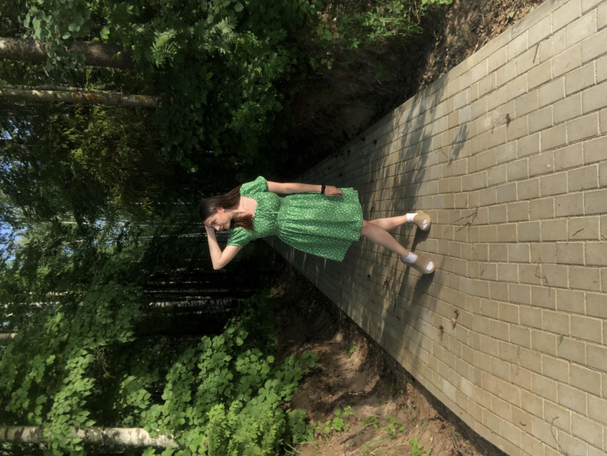 A young woman wearing a green dress is lying on a brick pathway, with trees and foliage on either side. The location is Noviy, Russia. She is resting her head on her hand.