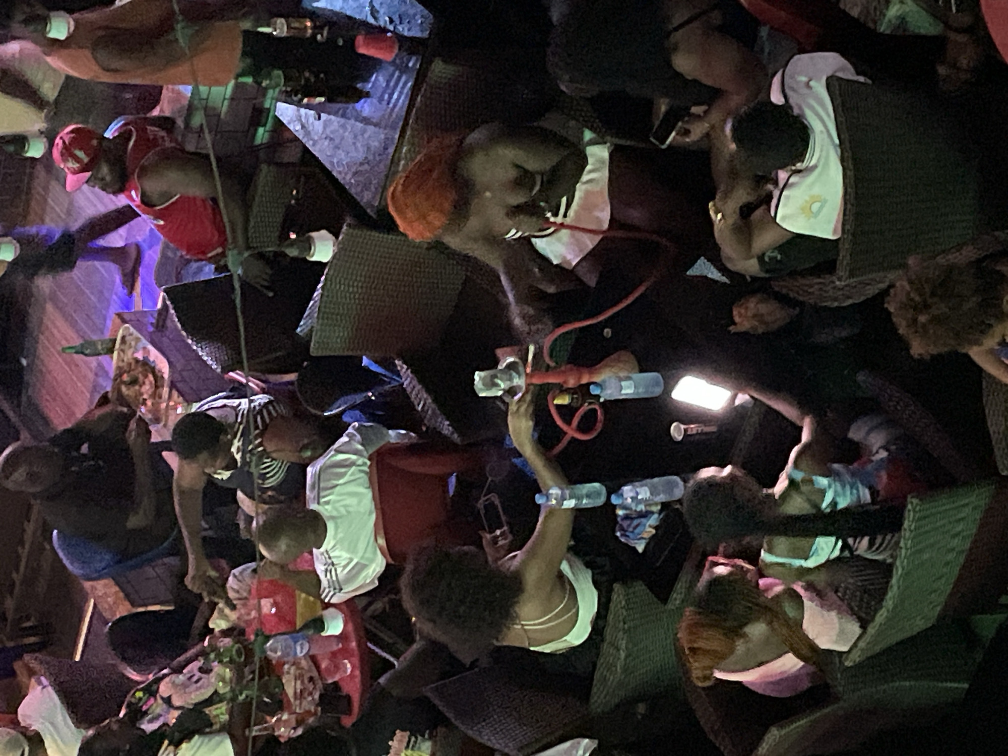 An aerial, medium shot captures a dimly lit, outdoor seating area at a nighttime establishment, possibly a bar or restaurant. Several individuals are seated at tables, some engaged in conversations or observing their surroundings. In the foreground, a woman with dark, curly hair is seated, holding a smartphone. To her right, another woman with bright orange hair is seated, holding a shisha pipe. On a table between them, a shisha apparatus is visible, along with a bottle of water and other items. The environment is furnished with woven chairs and tables, illuminated by ambient lighting that creates shadows. In the background, other patrons are visible, some standing and others seated, with food and drinks on their tables. The scene suggests a casual social gathering.