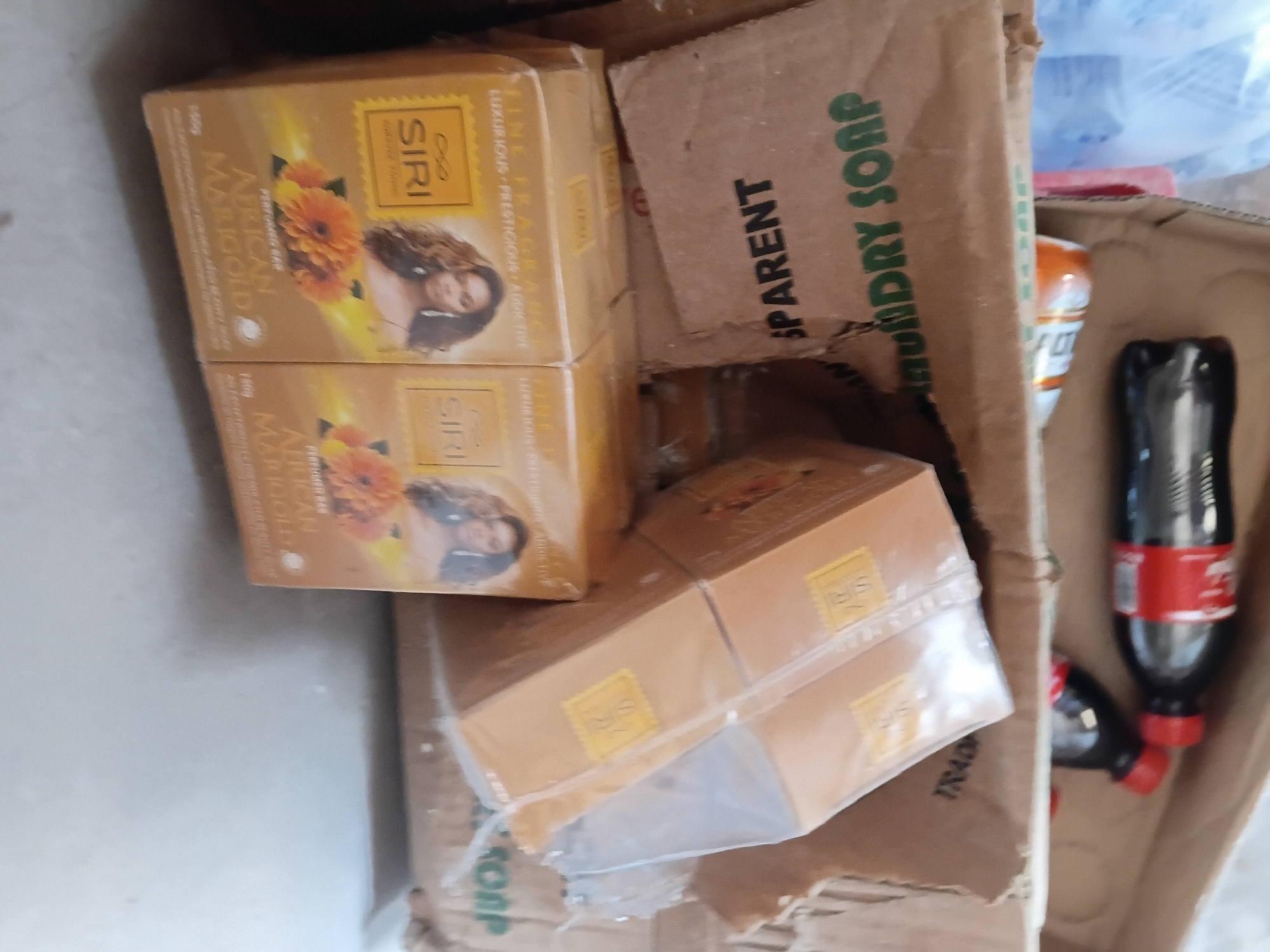 The image shows a close-up, overhead view of several cardboard boxes and some bottles of soda. In the foreground, two bottles of Coca-Cola are visible, one of which is partially obscured by the other. Behind these, there are several stacked cardboard boxes. The top boxes are branded with "SIRI AFRICAN MARIGOLD PERFUMED SOAP" and feature images of marigolds and a woman. Below these, another box is visible with the text "TRANSPARENT LAUNDRY SOAP" printed on it. The scene appears to be an informal storage area, possibly a shop or a warehouse, located in Jalingo, Nigeria. The lighting is moderate, and the overall impression is one of clutter and everyday commerce.
