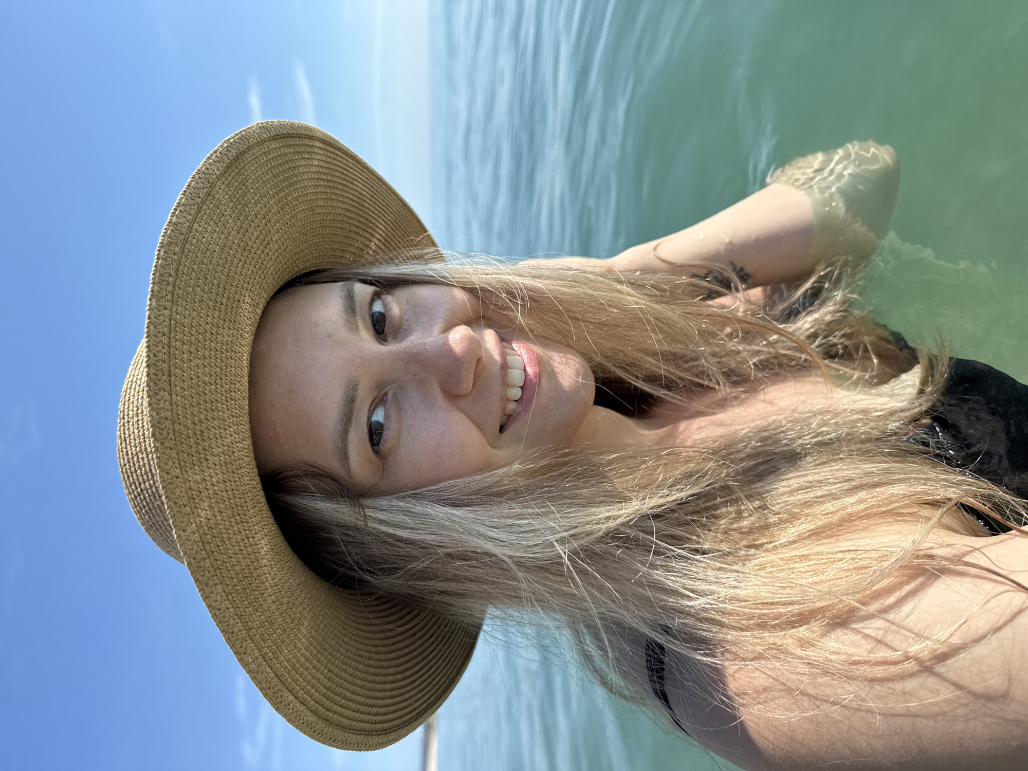 The image is a close-up selfie of a young Caucasian woman with blonde hair, wearing a straw hat and a black swimsuit, standing in the water. She is smiling and looking at the camera. The background shows a clear blue sky and the calm sea. The water is a light turquoise color. The scene appears to be a sunny day at the beach, possibly in the resort town of Nebug, Russia, given the context. The woman's left arm is visible, partially submerged in the water, and a faint tattoo is visible on her upper arm. The lighting suggests it is daytime, and the sun is high in the sky, casting soft shadows.