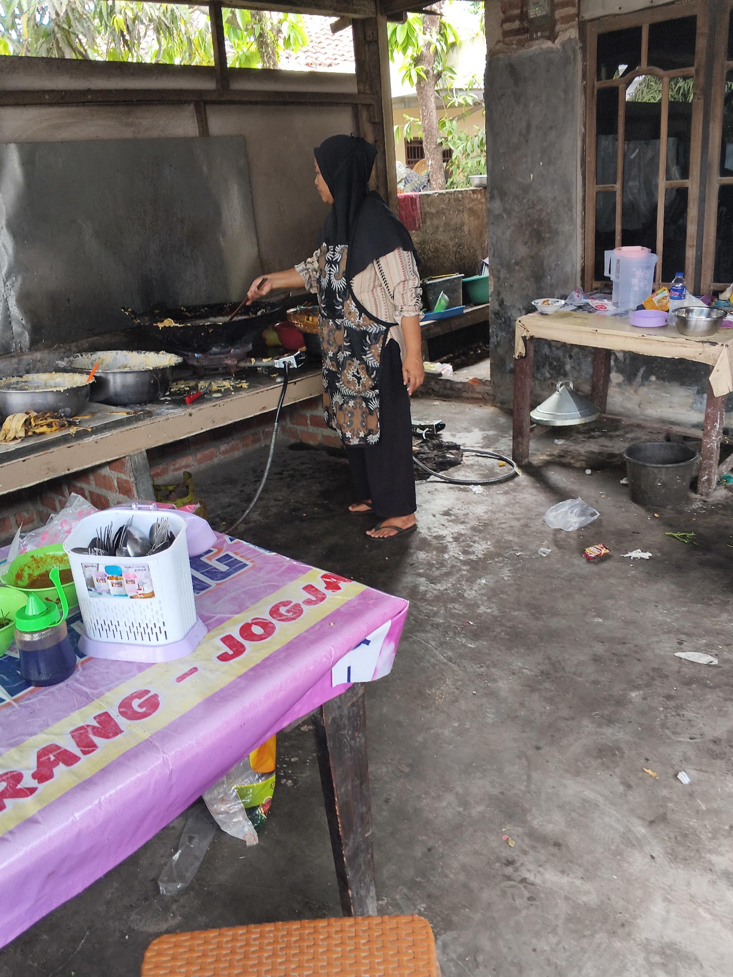The image shows a woman wearing a black hijab and a patterned apron, cooking at a wok on a stove in an outdoor market stall in Bandar Lampung, Indonesia. The stall appears to be a food vendor, with various cooking utensils, pots, and ingredients visible. The woman is actively stirring food in the wok. In the foreground, a pink and yellow tablecloth covers a table, with a white utensil holder filled with spoons and forks. The ground is concrete and appears somewhat dirty, with some debris scattered around. In the background, there is a wooden structure and some foliage, suggesting a tropical environment. The scene depicts a typical street food vendor operation in Indonesia.