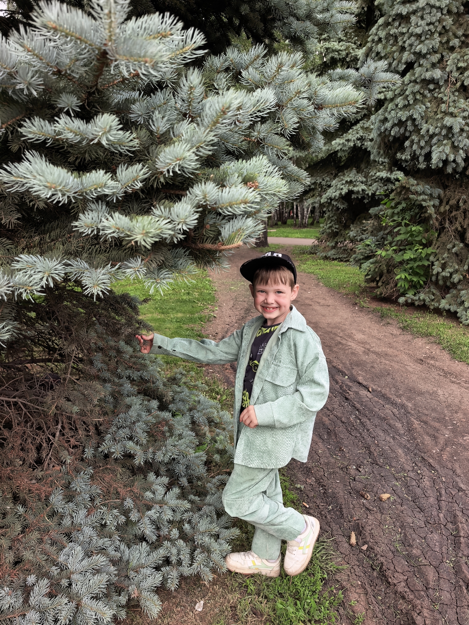 A young boy with a wide smile is posing next to a large fir tree on a dirt path in a park. He is wearing a light green corduroy suit, a black baseball cap, and white sneakers. The boy is looking at the camera and has his left hand extended towards the tree. The background shows more fir trees and green grass. The image appears to be taken outdoors in a park environment.
