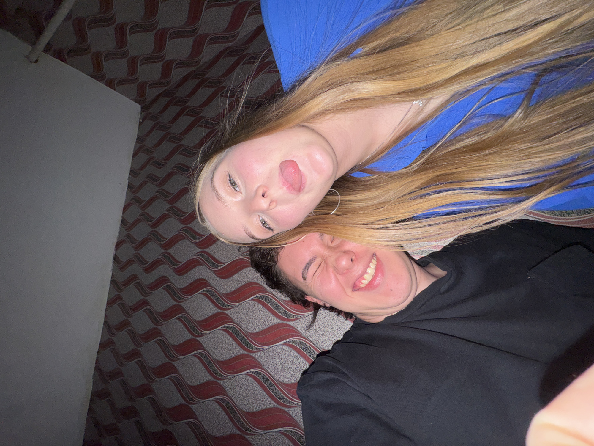 A low-angle selfie is taken indoors, with two young adults in the foreground. The person on the left, appearing male, is wearing a black t-shirt and has dark, curly hair. Their eyes are closed, and they are smiling broadly, showing their teeth. The person on the right, appearing female, has long, blonde hair and is wearing a bright blue top. Her tongue is sticking out, and she is looking directly at the camera. They are positioned close together, suggesting familiarity or a shared moment. Behind them, the wall is covered in a distinctive red and grey patterned wallpaper, featuring wavy vertical stripes. The ceiling above is a plain, light color. The lighting appears to be artificial, creating shadows and highlighting the subjects. The overall impression is one of a casual and playful interaction. The location is identified as being in the city of Perm, Russia.