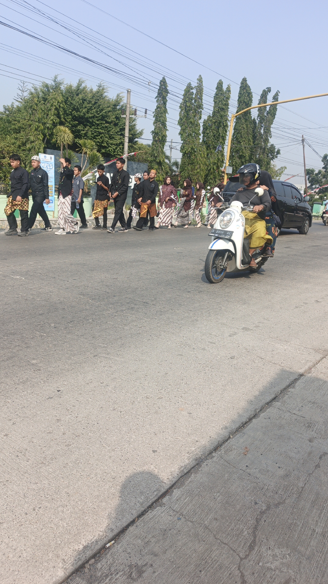 In Sumengko, Indonesia, a group of young people, predominantly male and wearing traditional attire including black shirts and patterned sarongs, walk along a paved street under a clear sky. To the right, a person rides a white scooter, also wearing traditional clothing and a helmet, with another person on the back of the scooter. A black car is visible behind the scooter, and in the background, a line of tall, slender trees and utility poles are present. The street appears to be a road in a town or city, with concrete sidewalks and asphalt pavement.
