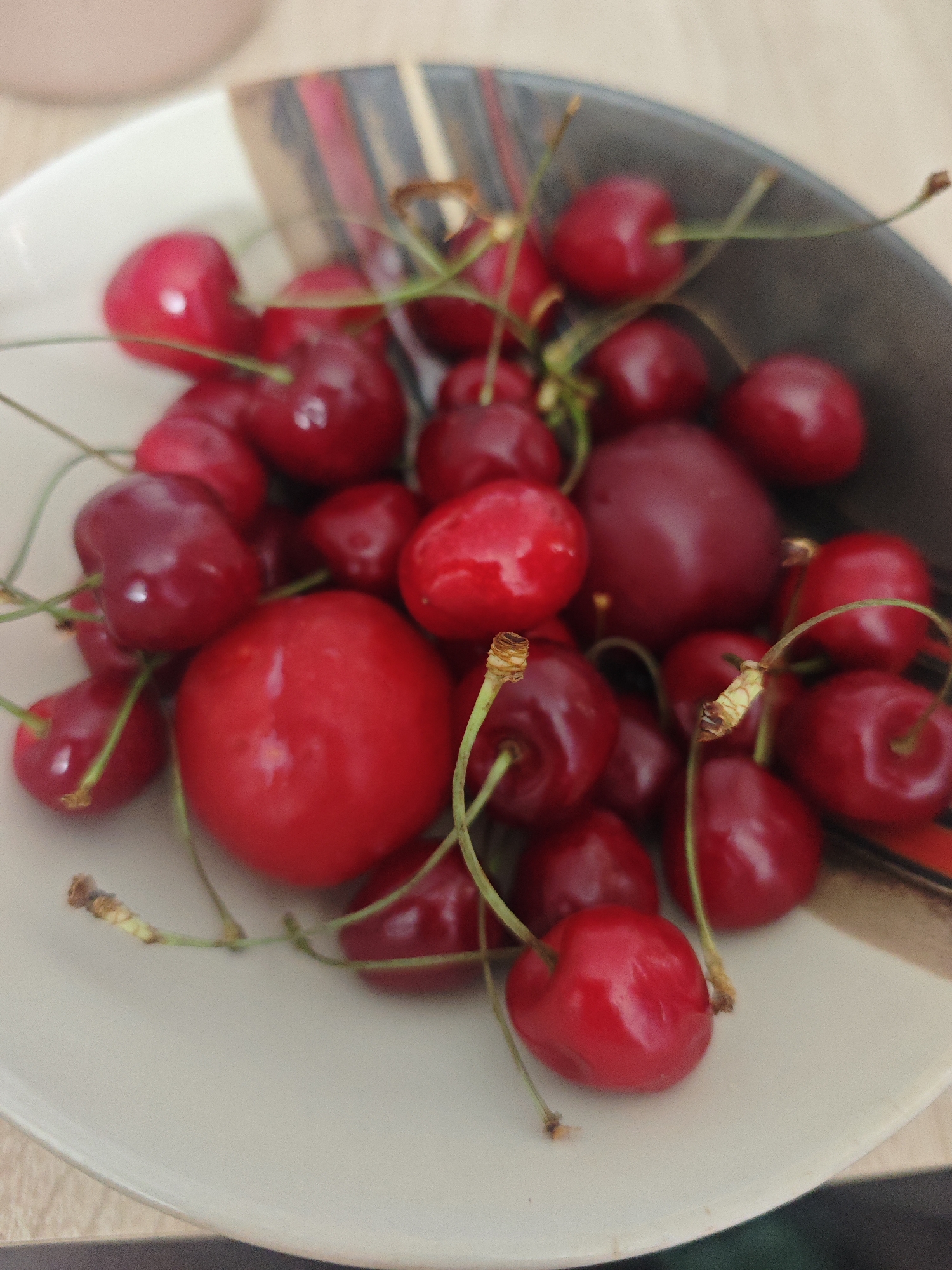 A close-up, overhead shot showcases a bowl filled with ripe red cherries. The cherries, some with stems, are clustered together, creating a vibrant visual. The bowl itself has a textured, dark gray rim with subtle red and black stripes. The background is blurred, suggesting an indoor setting with a light-colored wooden surface beneath the bowl. The image is sharp and focused on the cherries. The scene is devoid of any people or specific activities. There is no information in the image to suggest the location in the city of Chekhov, Russia.