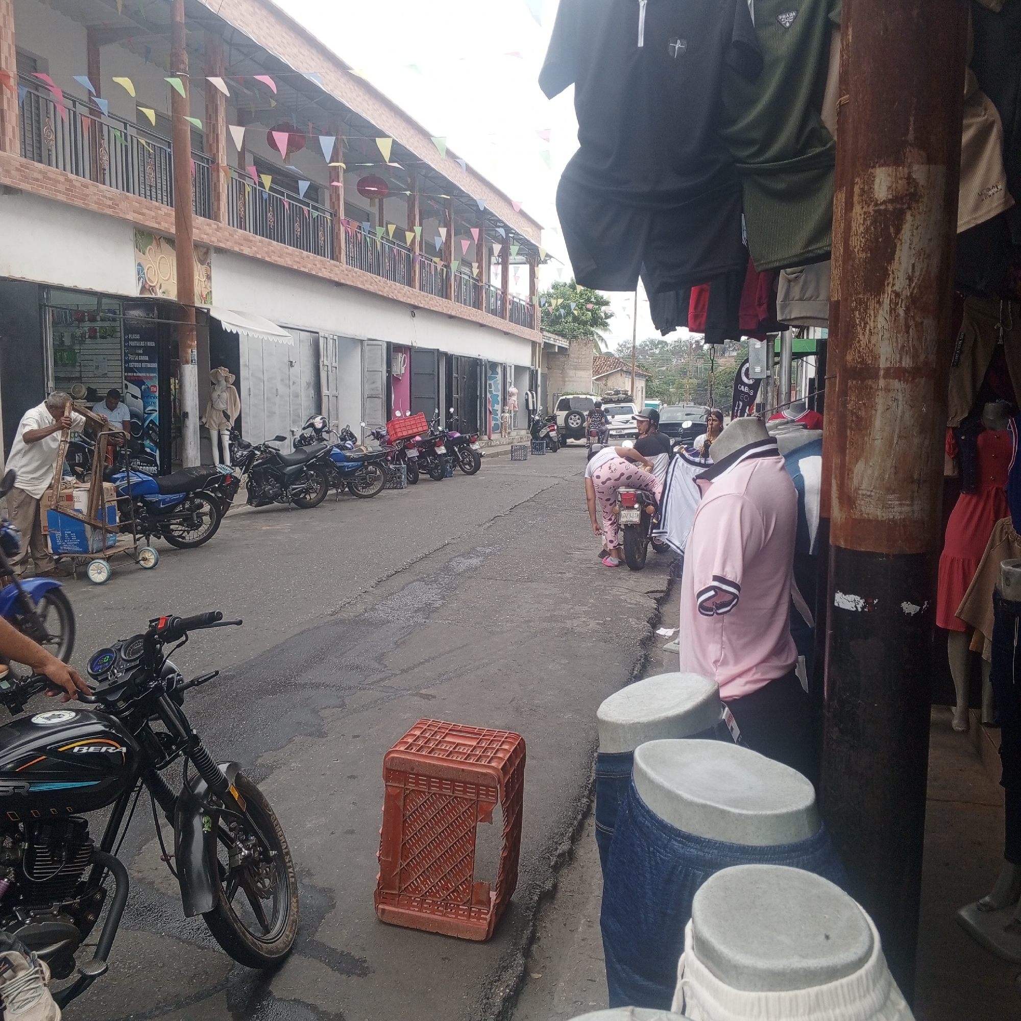 The image captures a street scene in Tinaquillo, Venezuela. In the foreground, a black Bera motorcycle is parked. Next to it, a damaged orange plastic crate rests on the cracked asphalt. To the right, mannequin torsos wearing shorts and pants stand on display outside a store. A rusty metal pole is positioned nearby, with clothing hanging from it, obscuring part of the scene.
In the middle ground, a woman in pink pajamas attends to a motorcycle, while another person sits on it. Further down the street, multiple motorcycles are parked along the left side of the road, near businesses. A man is seen pushing a cart loaded with items. In the background, cars and people are visible. The street is lined with two-story buildings adorned with colorful pennant banners.