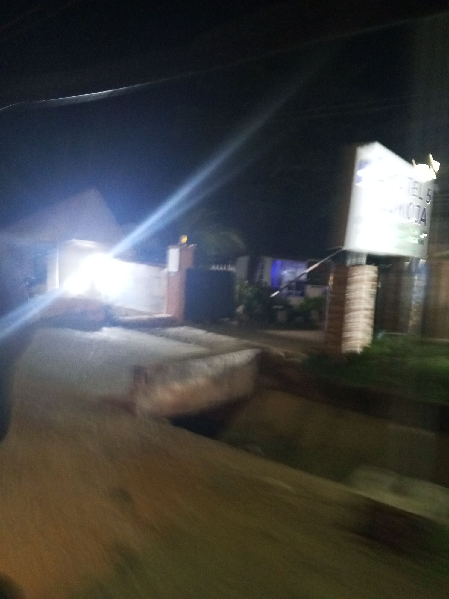 The image, taken at night in Lokoja, Nigeria, shows a dimly lit street scene. A brightly lit structure, possibly a building or a shop, is visible on the left with a powerful light source illuminating the area. In the foreground, a road or pathway with a slightly elevated curb is present. To the right, a tall signpost with the text "TEL 5 MUOJA" is illuminated, supported by brick pillars. Further in the background, other buildings and vegetation can be discerned amidst the darkness. The overall impression is of a roadside establishment in an urban or semi-urban setting at night.