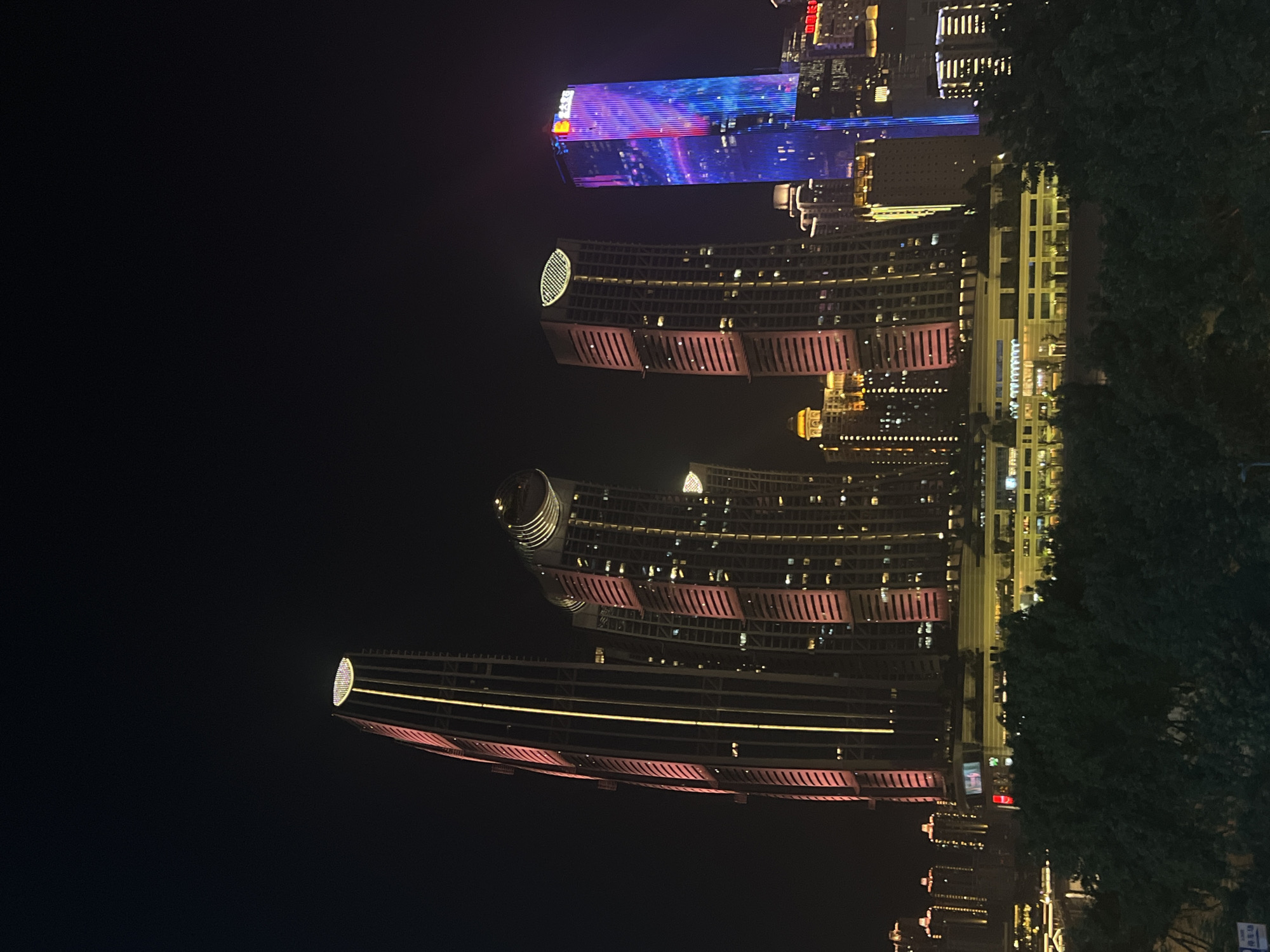 The image displays a night scene of a modern cityscape, likely in Jiangbeicheng Street, China, characterized by illuminated skyscrapers. The prominent structures are tall buildings with diverse architectural designs, showcasing an array of lights in various colors and patterns. One very tall skyscraper on the left features vertical yellow and pink lighting, with a circular, illuminated top. To its right, a cluster of buildings with similar vertical lighting are visible, some with rounded or segmented tops. Further to the right, a particularly striking skyscraper is adorned with vibrant blue and pink vertical light displays, creating a dynamic visual effect. Smaller buildings with horizontal and vertical lights are scattered throughout the scene, some appearing as office buildings or apartments. In the foreground, a dense canopy of dark green trees creates a natural border, obscuring the lower parts of the urban landscape. The overall impression is one of a bustling, technologically advanced city at night, with the buildings acting as focal points against the dark sky. There are no people or specific actions depicted in the image, and the environment is dominated by artificial illumination from the architecture.