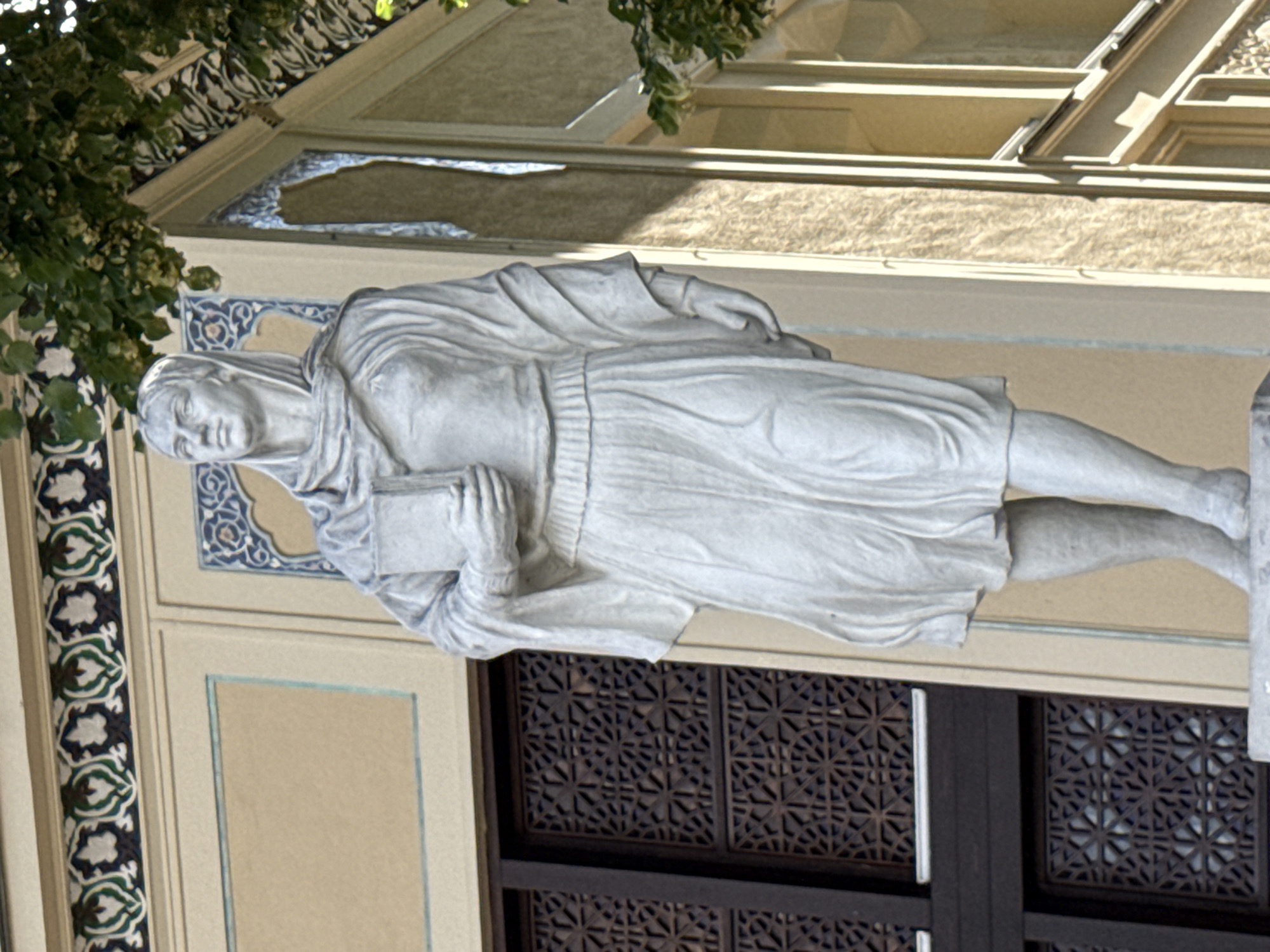The image shows a stone statue of a woman holding a book in front of a building. The woman is depicted wearing a draped robe and a headscarf. The building has architectural elements that appear to be in a Moorish or Islamic style, with decorative tile work and arched window frames. The statue is positioned on a raised platform. The background shows part of a window with a decorative lattice and trees with green foliage overhead. This statue is located in Moscow, Russia, specifically on the facade of the Moskva Museum of Modern Art, which was formerly the headquarters of the National Bank of the Russian Empire.