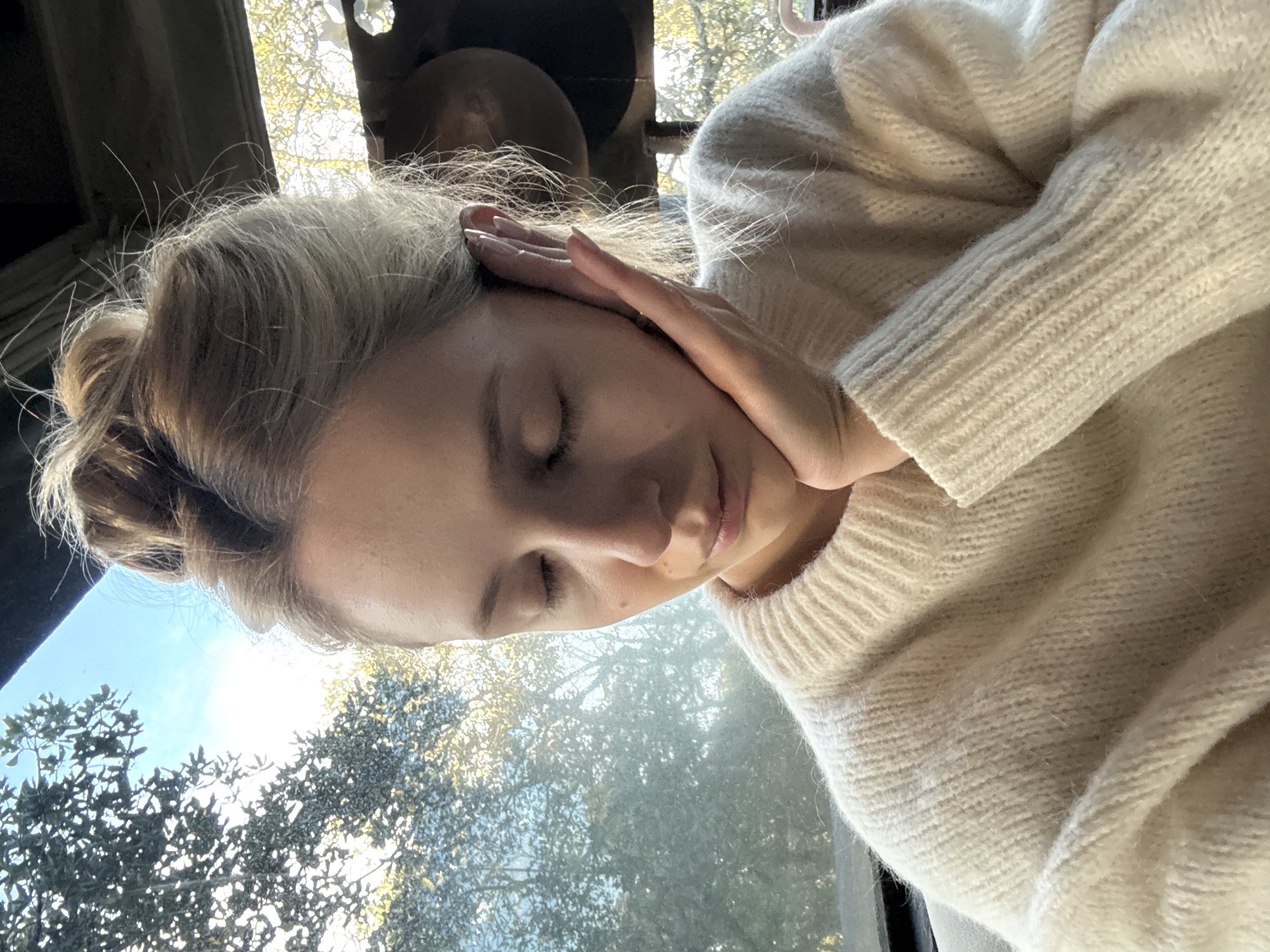 A medium shot captures a young Caucasian woman with blonde hair pulled up in a messy bun, her eyes closed, and her right hand resting on her cheek as she leans her head. She is wearing a cream-colored, chunky knit sweater. The background is blurred, showing trees and a bright sky through a window, suggesting an outdoor or well-lit indoor setting. There is no discernible indication of the location within Ciudad de México, Mexico.