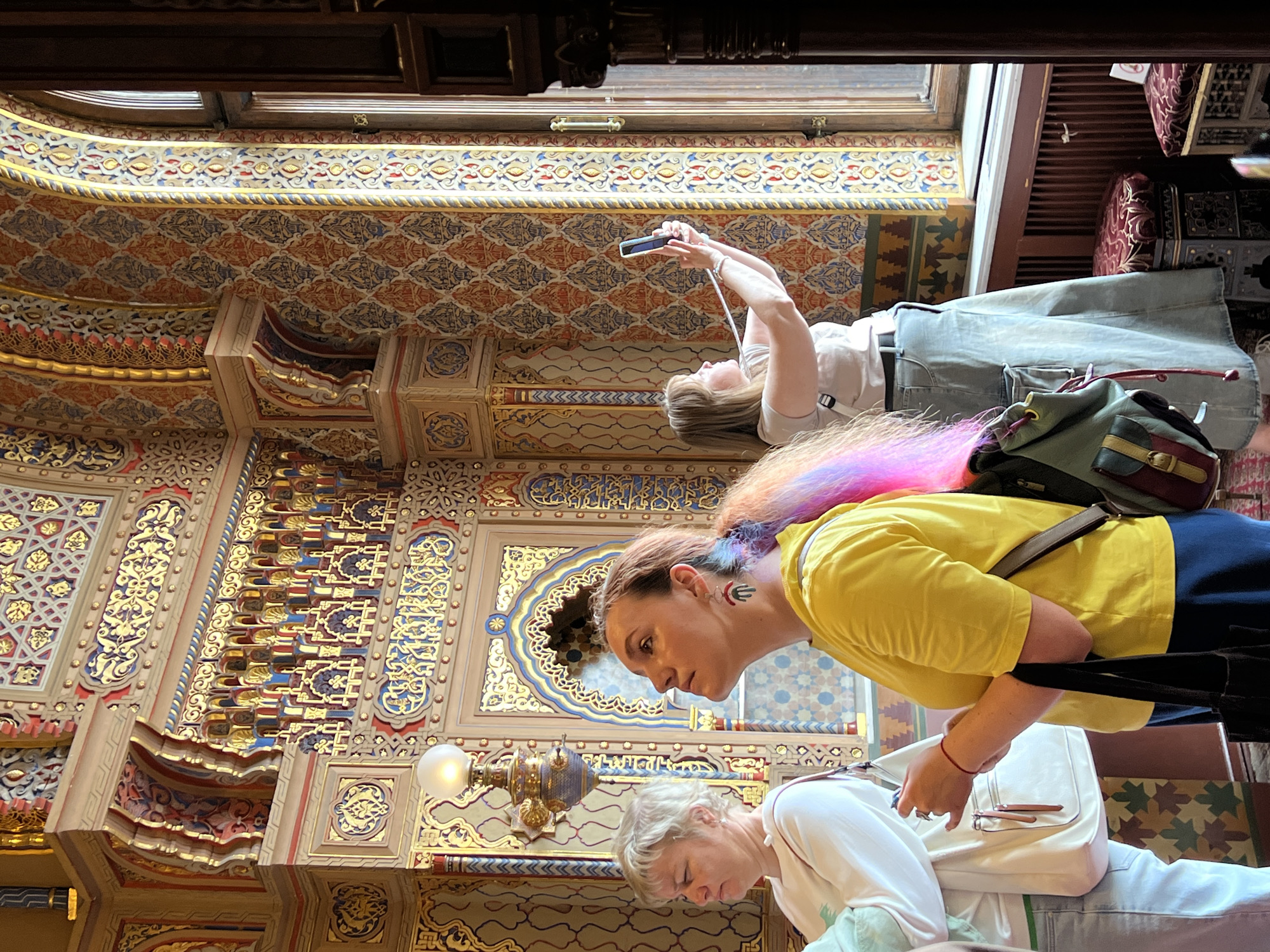 In Saint Petersburg, Russia, inside a room adorned with intricate, colorful patterns and golden embellishments on the walls, three women are present. The woman on the left, with short blonde hair, is wearing a white long-sleeved shirt and light-colored pants, carrying a cream-colored bag over her shoulder. In the center, a woman with a multi-colored ponytail is looking upwards, dressed in a bright yellow t-shirt and dark blue skirt, holding a black tote bag. The woman on the right, with blonde hair, is taking a photo with her smartphone, wearing a light-colored shirt and a long denim skirt, and has a white strap from a bag around her neck. The room features a tall arched window, and ornate architectural details are visible throughout.